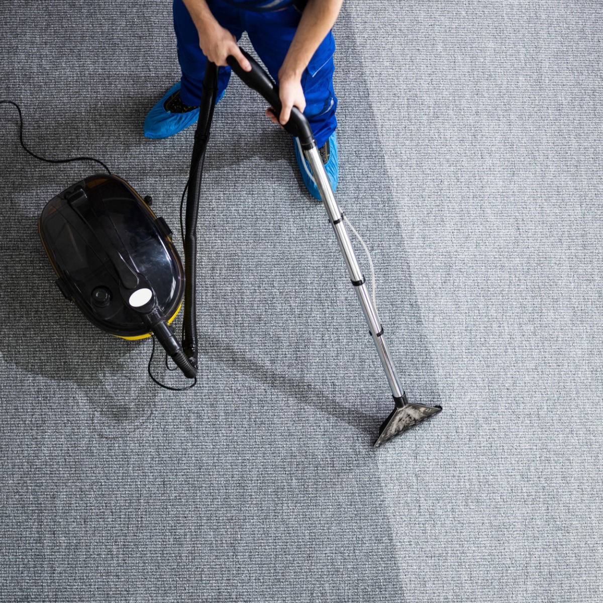 Widok z góry na pracownika w niebieskim kombinezonie czyszczącego szary dywan za pomocą odkurzacza piorącego, widoczny efekt czyszczenia.
