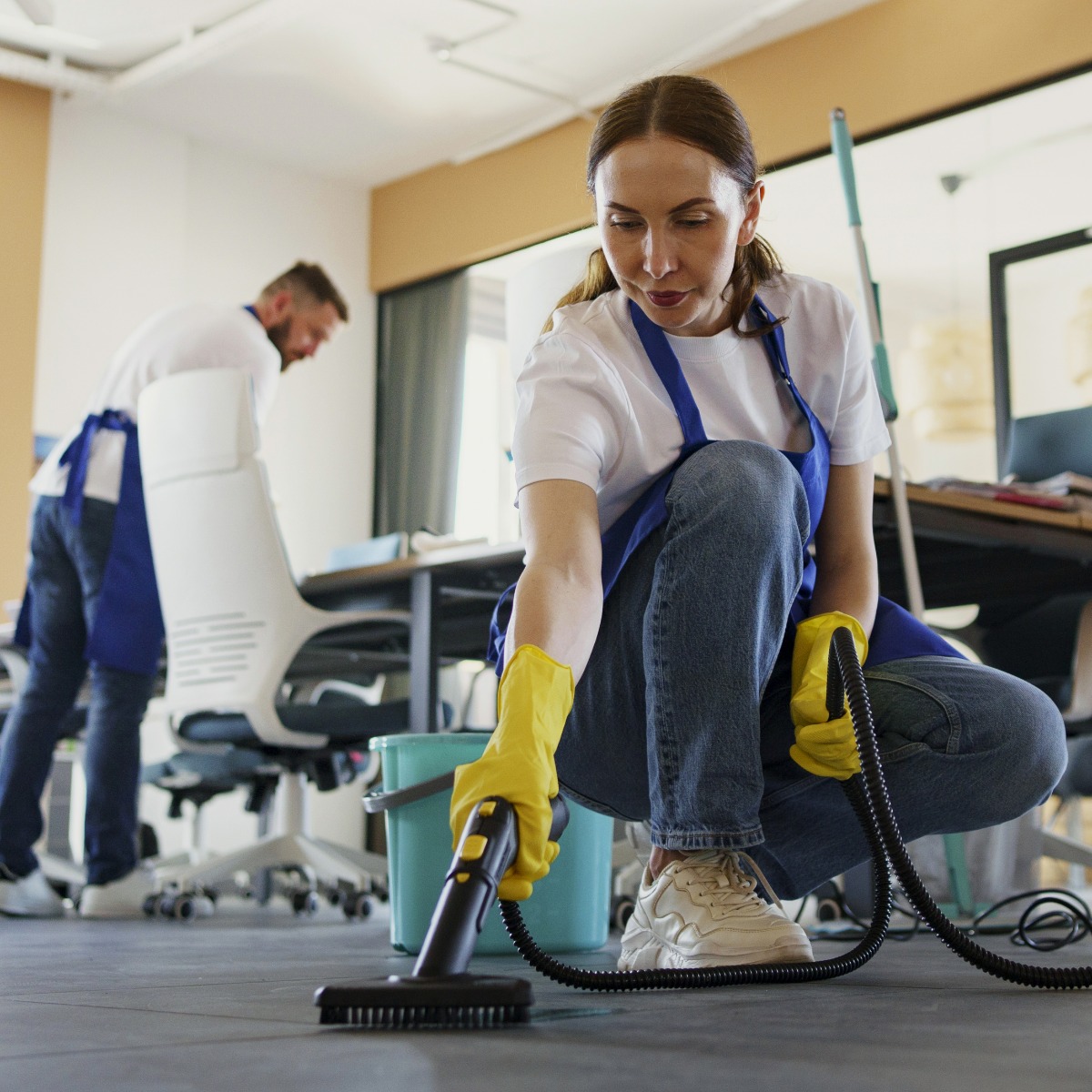 Kobieta w niebieskim fartuchu i żółtych rękawicach czyści podłogę w biurze za pomocą odkurzacza z szczotką, obok stoi wiadro, w tle widoczny mężczyzna sprzątający biurko.