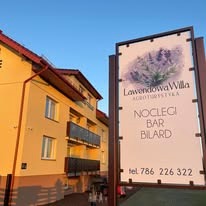 Tablica reklamowa przed budynkiem z napisem 'Lawendowa Willa AGROTURYSTYKA', oferująca noclegi, bar i bilard, z grafiką lawendy w górnej części.