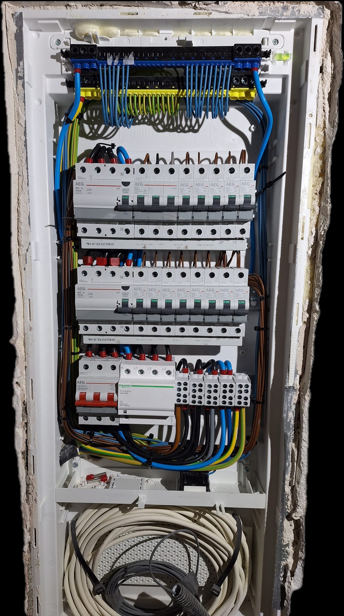 Rozdzielnica elektryczna z zainstalowanymi bezpiecznikami i okablowaniem w białej obudowie, widoczne marki AEG i Schneider, ściana wokół obudowy nierówna i otynkowana