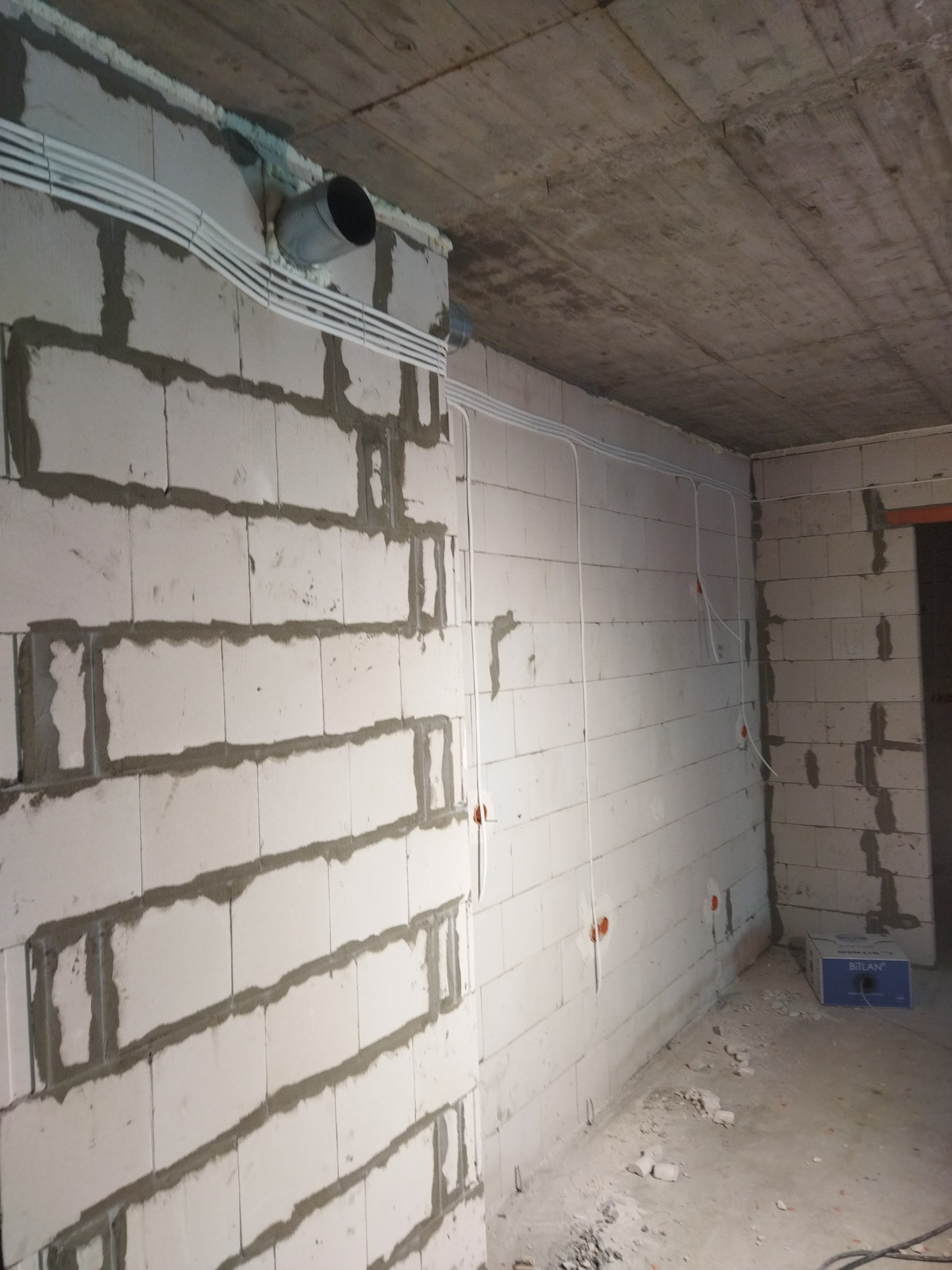 Surowy betonowy sufit i ściany z bloków betonowych w trakcie budowy, widoczne białe rury instalacji elektrycznej prowadzone wzdłuż ścian i pod sufitem, oraz króciec wentylacyjny przechodzący przez...