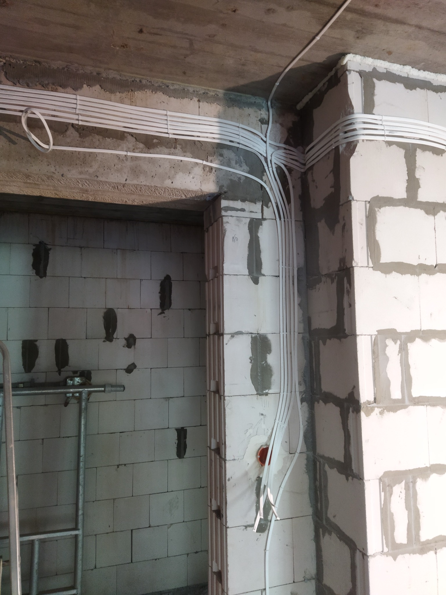 Białe rury instalacji elektrycznej ułożone równolegle na surowym suficie i ścianie z białych bloczków, widoczne puszki instalacyjne i drabina w tle.