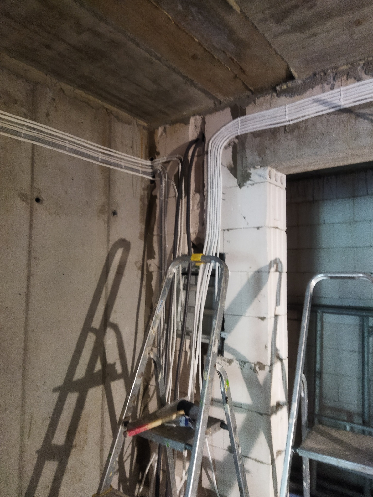 Białe kable elektryczne prowadzone wzdłuż surowej ściany z betonu komórkowego, widoczna drabina z młotkiem na półce, surowe wykończenie wnętrza.
