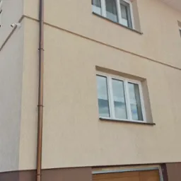 Dwukondygnacyjny dom z beżową elewacją, brązowym cokołem i brązową rynną spustową, widok z dołu na okna i bramę garażową.