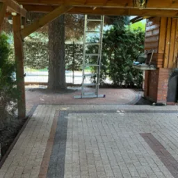 Ułożona kostka brukowa w odcieniach szarości i brązu pod drewnianą konstrukcją z widoczną drabiną aluminiową i drzewem w tle.