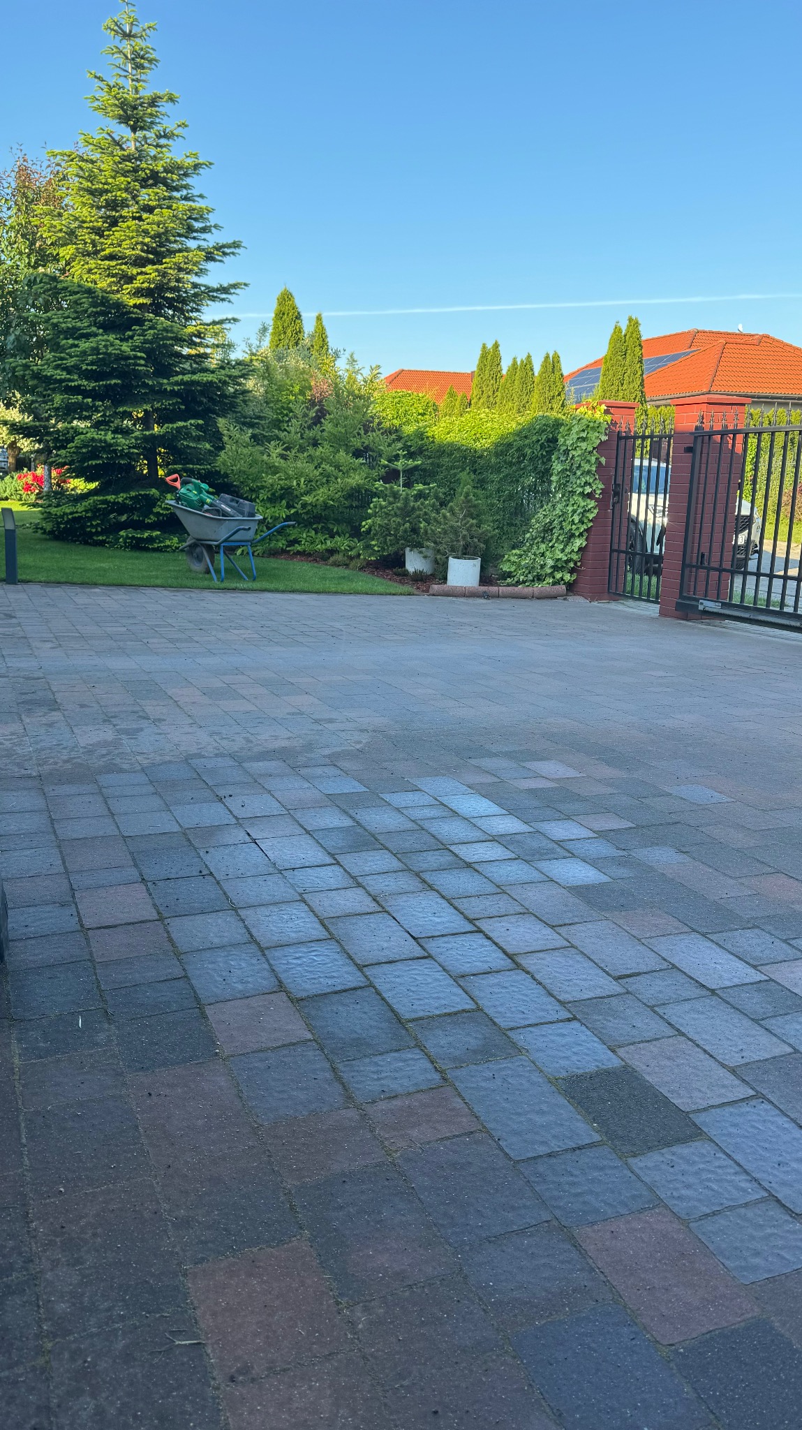 Częściowo umyta kostka brukowa na podjeździe, widoczna różnica w kolorze, taczka z narzędziami ogrodniczymi w tle.