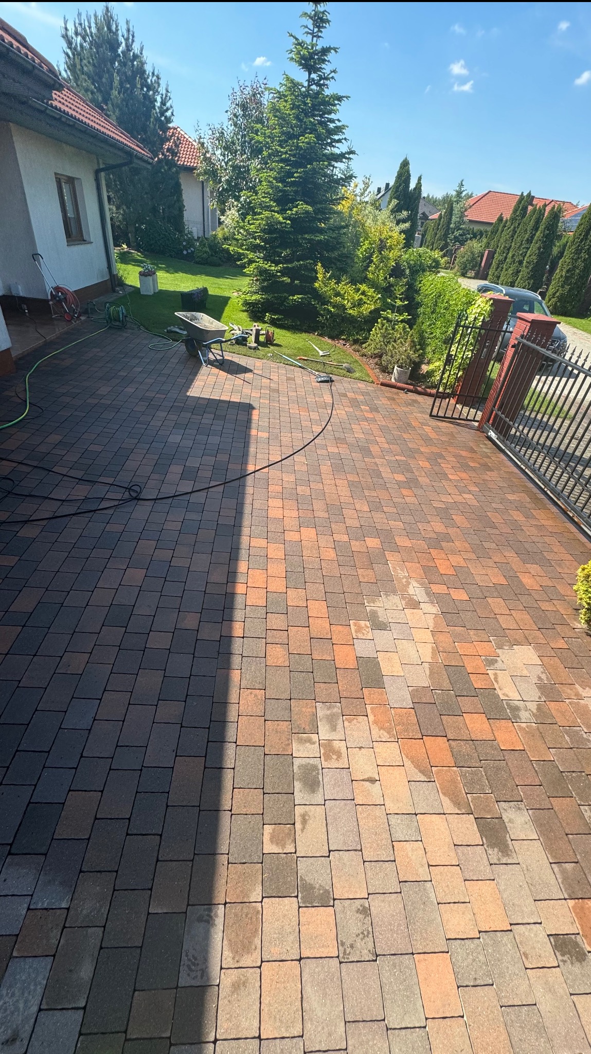 Częściowo umyta kostka brukowa w odcieniach brązu i szarości, widoczny wąż ciśnieniowy, taczka i narzędzia ogrodowe na trawniku w tle, brama wjazdowa po prawej stronie.