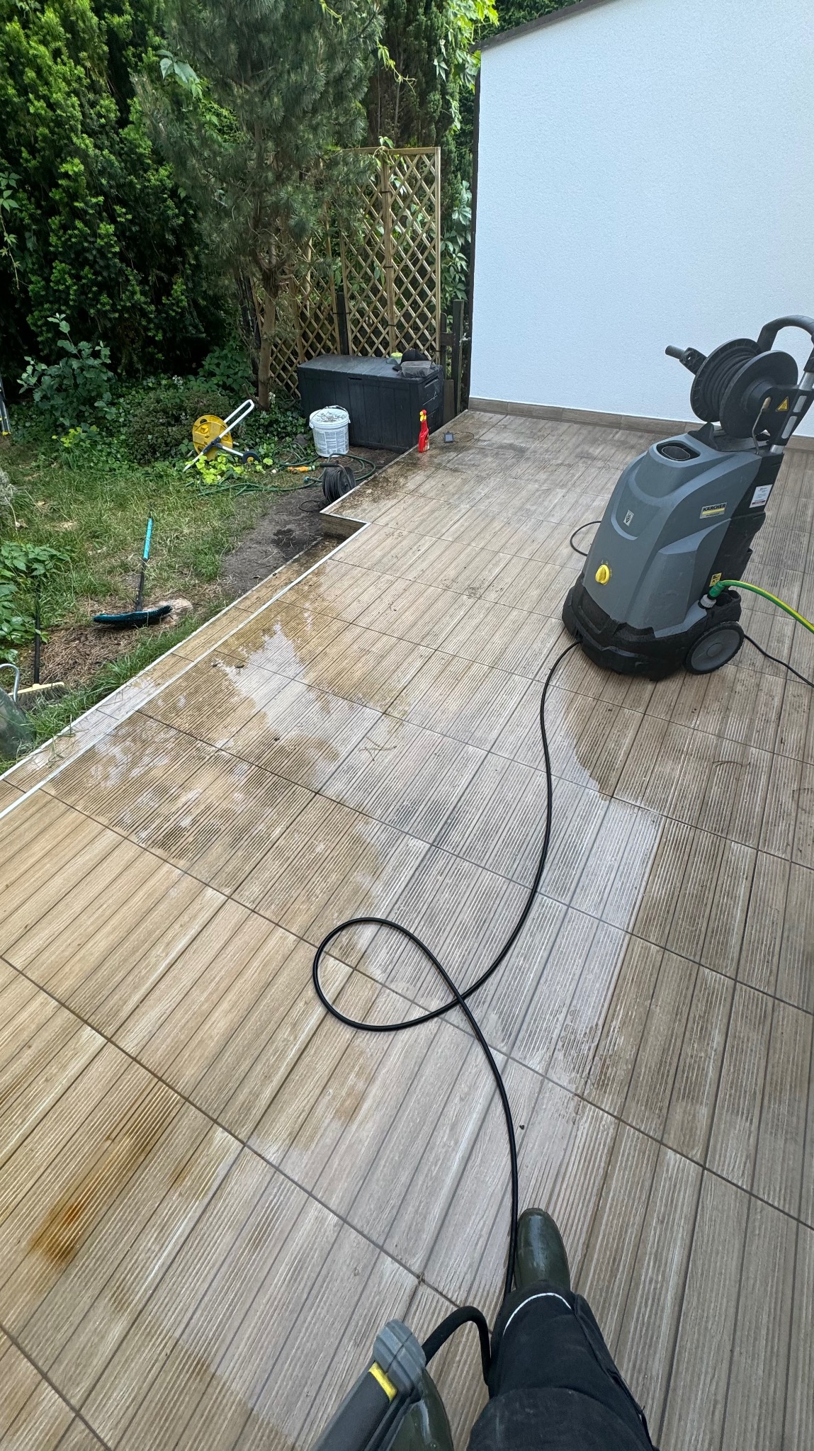 Czyszczenie tarasu z płytek ceramicznych przy użyciu myjki ciśnieniowej Karcher, widoczne mokre i czyste fragmenty oraz zabrudzenia przed czyszczeniem.
