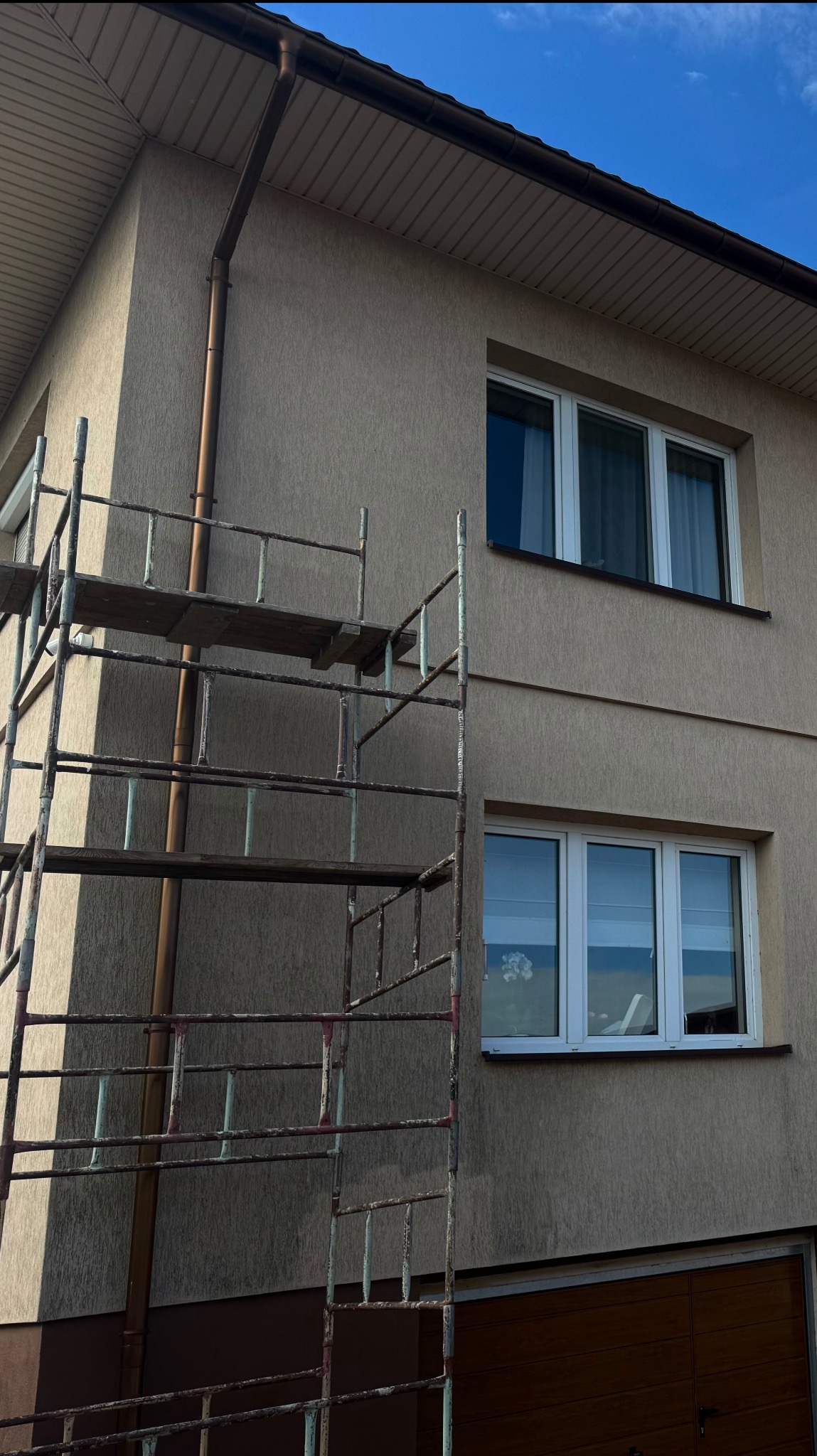Dom z beżową elewacją, okna z białymi ramami i metalowe rusztowanie przy ścianie, widoczny brązowy orynnowanie i błękitne niebo.