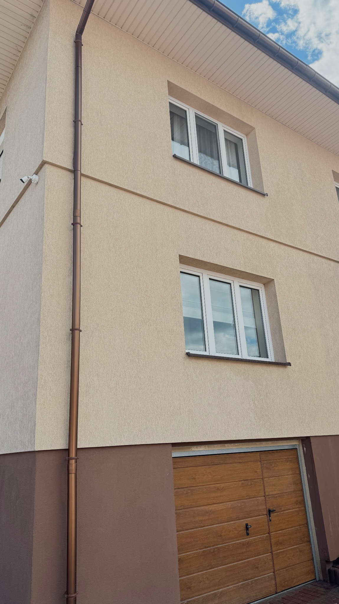 Dwukondygnacyjny dom z beżową elewacją, brązowym cokołem i brązową rynną spustową, widok z dołu na okna i bramę garażową.