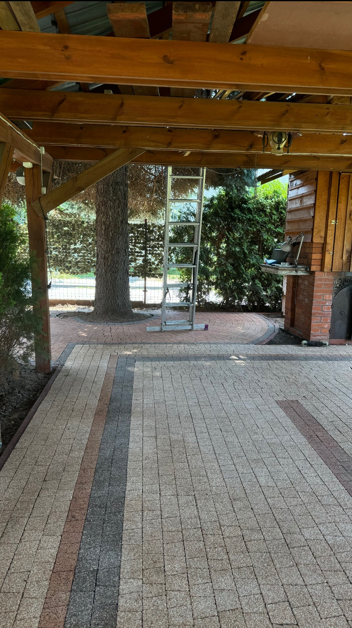 Ułożona kostka brukowa w odcieniach szarości i brązu pod drewnianą konstrukcją z widoczną drabiną aluminiową i drzewem w tle.