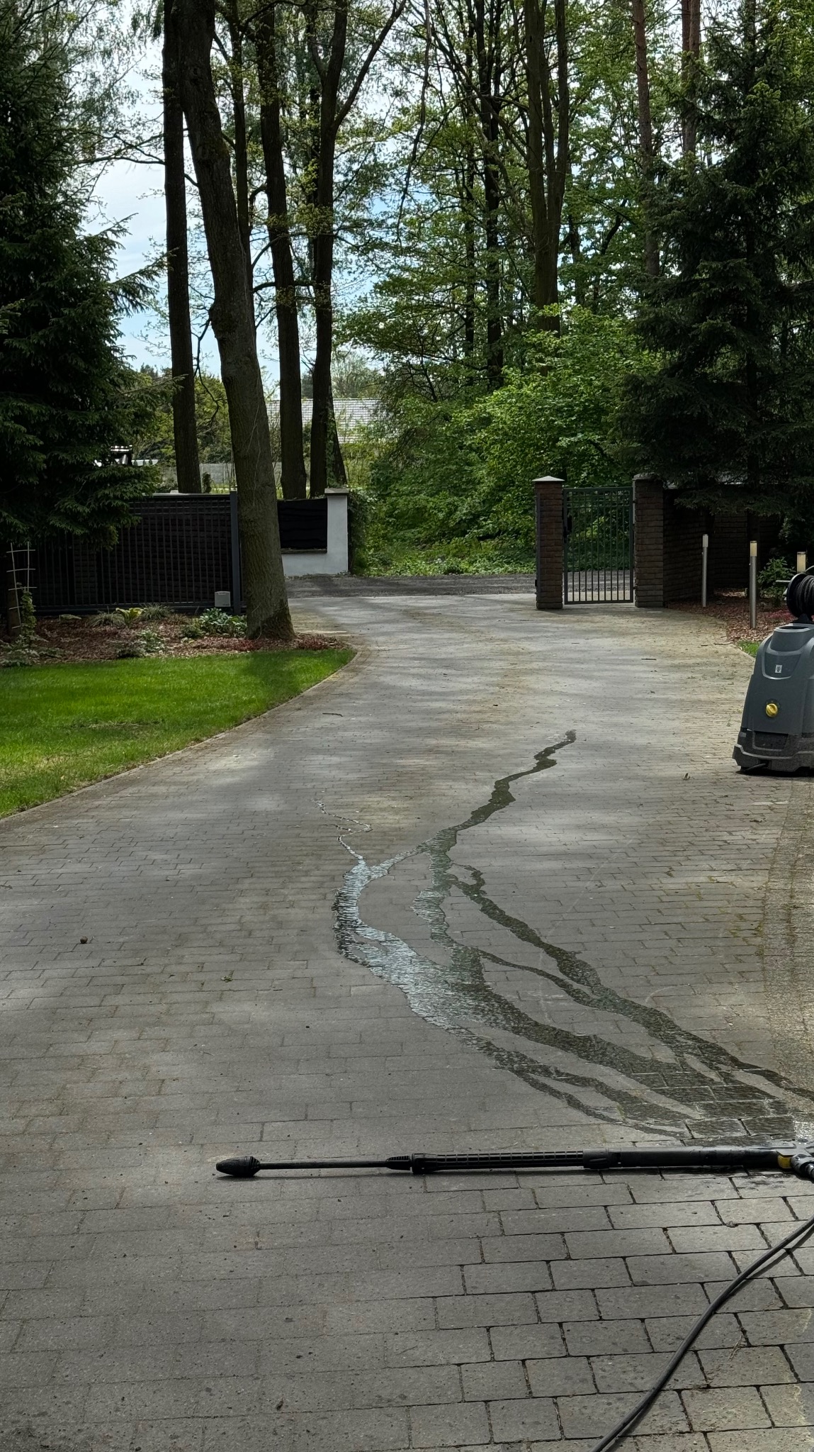 Częściowo umyta kostka brukowa na podjeździe, widoczne ślady wody po czyszczeniu, na pierwszym planie lanca myjki ciśnieniowej, w tle brama wjazdowa i zieleń drzew.