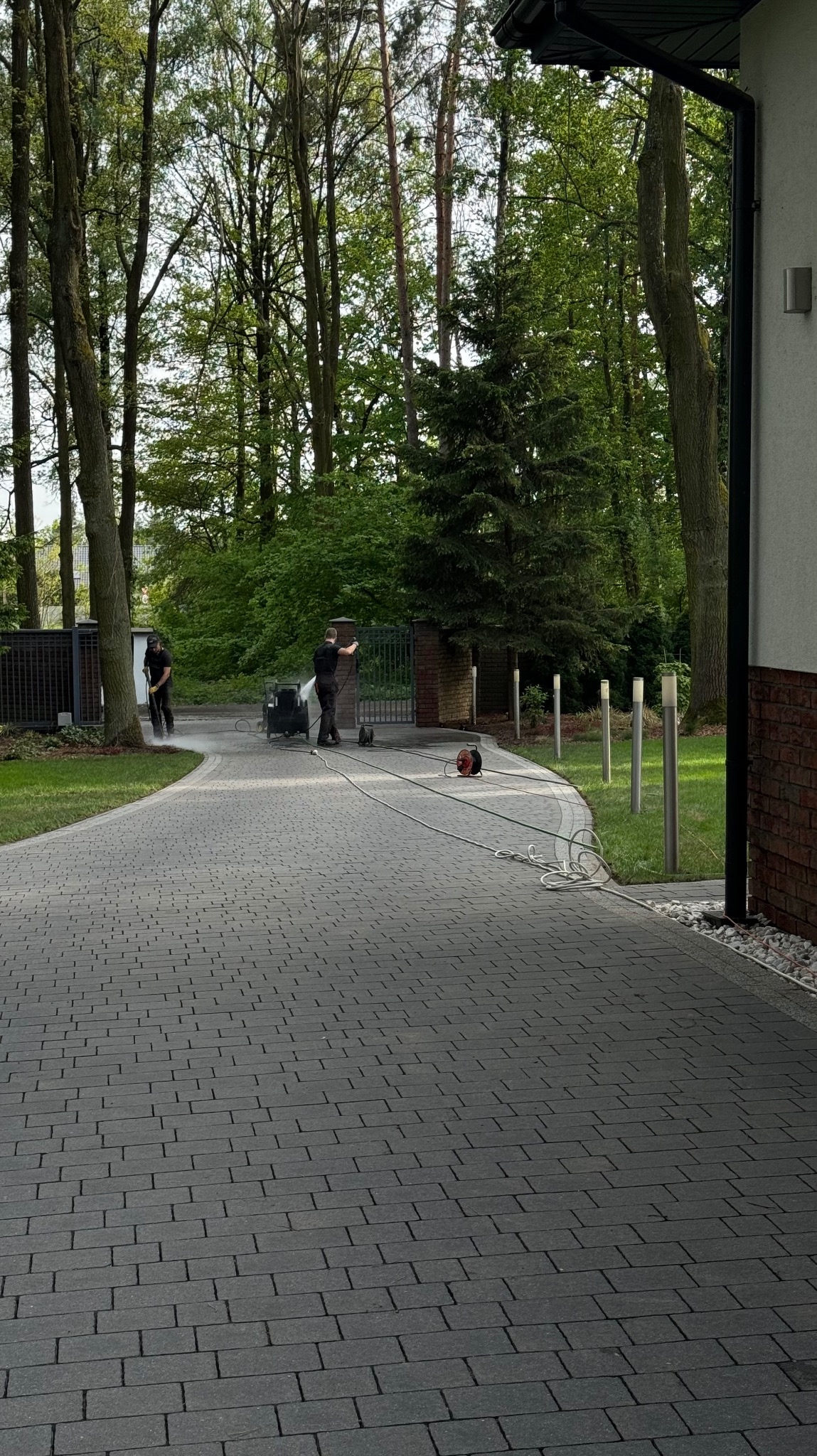 Dwóch mężczyzn w czarnych ubraniach czyści kostkę brukową na podjeździe za pomocą myjki ciśnieniowej. Widoczne drzewa w tle i elementy architektury domu.