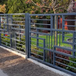 Nowoczesne przęsła ogrodzeniowe z metalu, pomalowane na szaro, z geometrycznym wzorem, oddzielające posesję od chodnika i rabaty kwiatowe