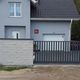 Nowoczesna, przesuwna brama wjazdowa z pionowymi, grafitowymi szczeblami, osadzona między słupkami z jasnoszarej kostki, na tle szarego domu z ciemnym dachem.