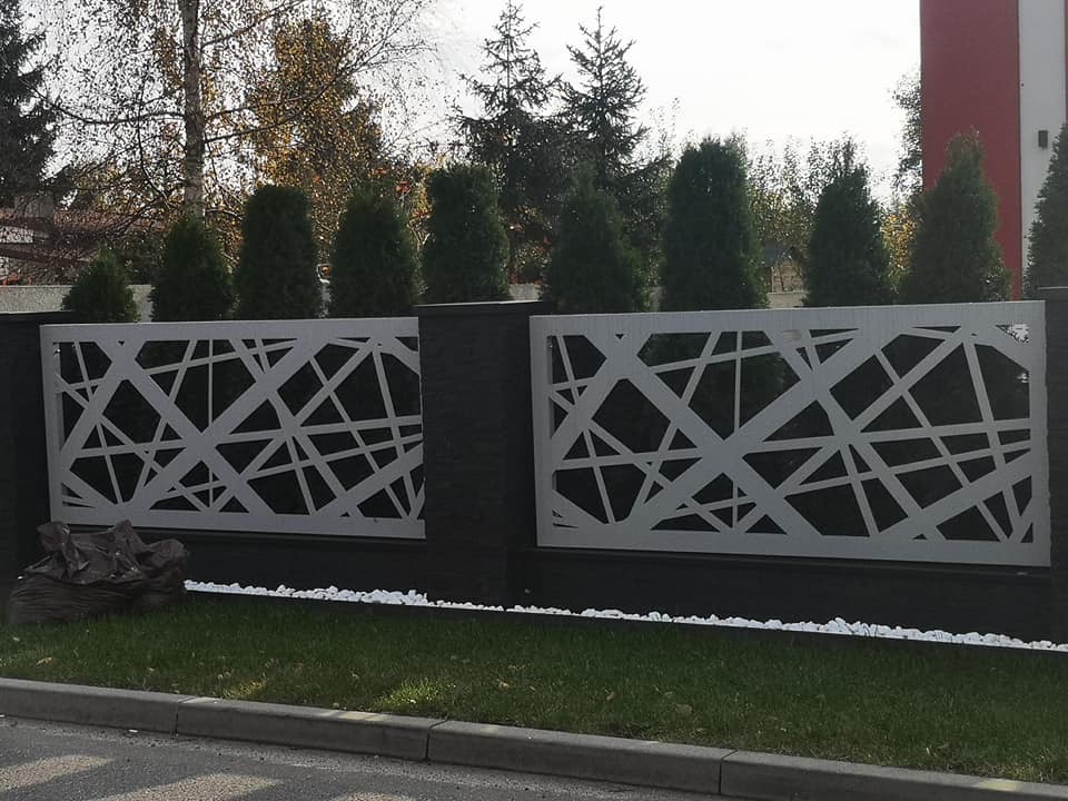 Nowoczesne ogrodzenie posesji z białymi, ażurowymi panelami o abstrakcyjnym wzorze na tle ciemnych słupków i zielonych drzew. Wzdłuż podstawy ogrodzenia ułożone białe kamienie.