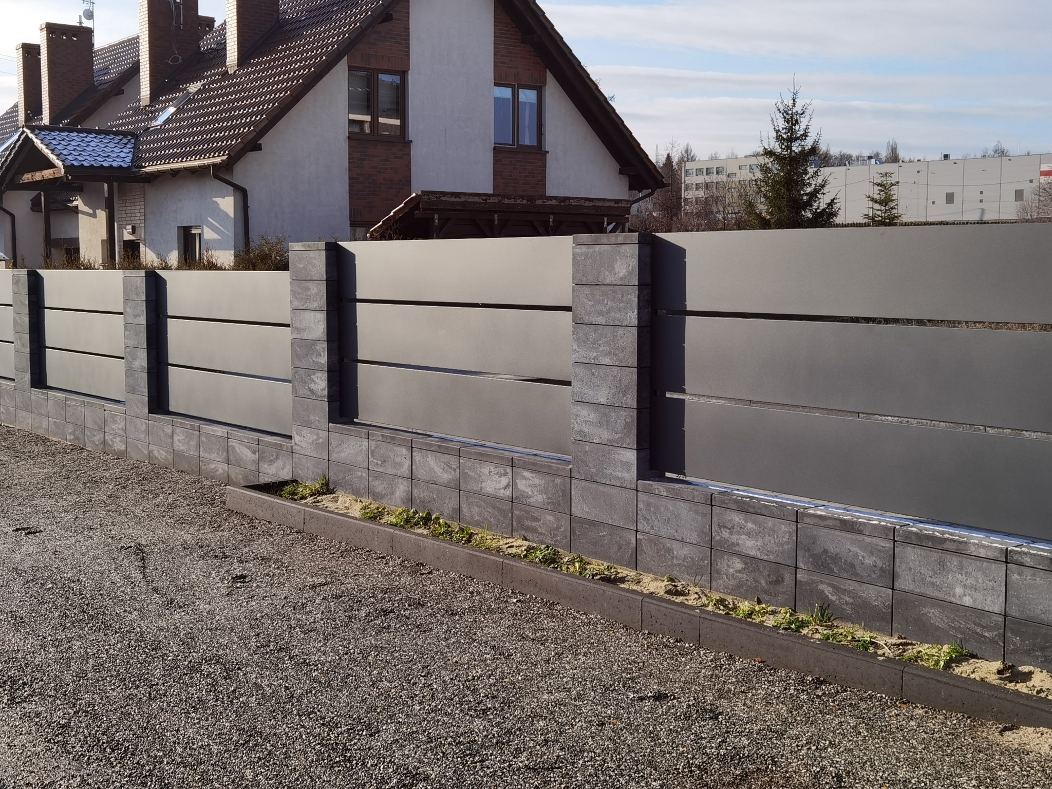 Nowoczesne ogrodzenie panelowe w kolorze grafitowym z elementami betonowymi, otoczone szarą nawierzchnią żwirową, na tle domu jednorodzinnego z brązową dachówką i budynkiem w oddali.