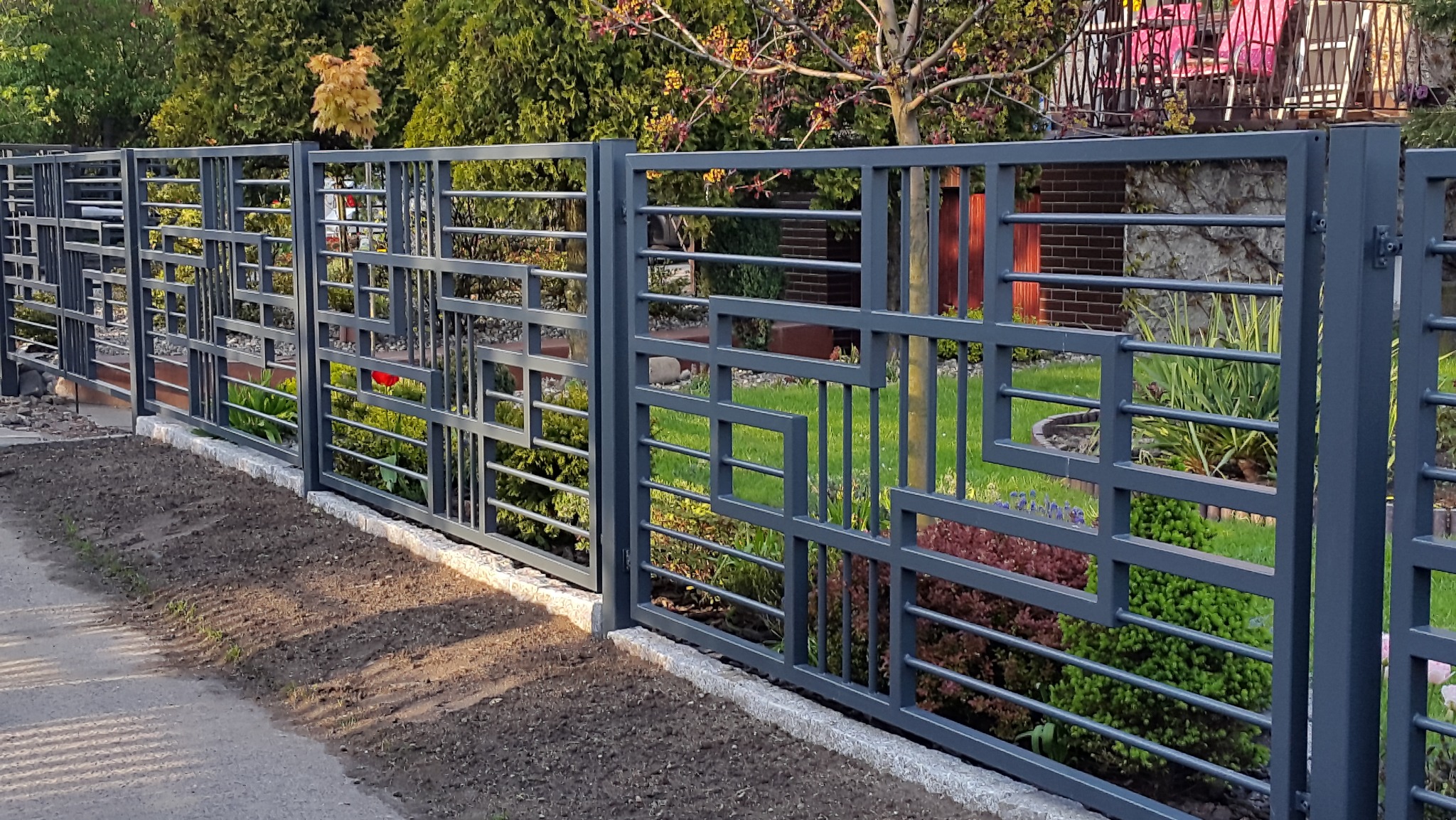 Nowoczesne przęsła ogrodzeniowe z metalu, pomalowane na szaro, z geometrycznym wzorem, oddzielające posesję od chodnika i rabaty kwiatowe