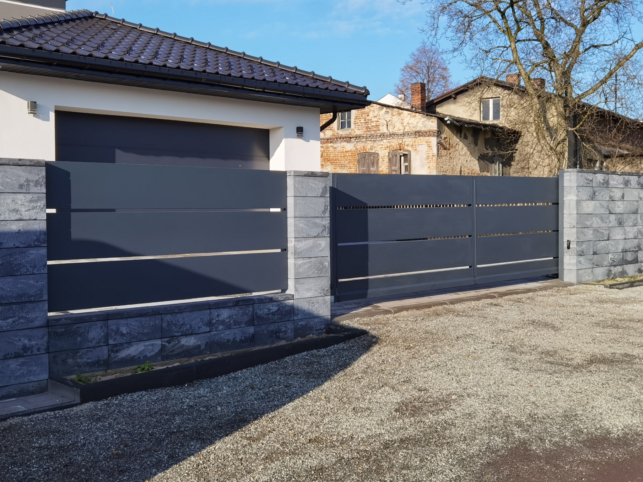 Nowoczesna, przesuwna brama wjazdowa z szarego metalu z poziomymi prześwitami, osadzona na murku z szarej kostki łupanej, na tle domu z garażem i starszego budynku.