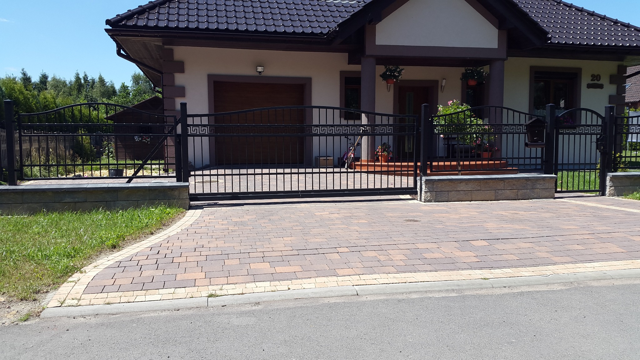 Czarna, metalowa brama wjazdowa z motywem greckim, zamontowana przed domem w Orzeszu, z brukowanym podjazdem i fragmentem ogrodzenia po bokach.