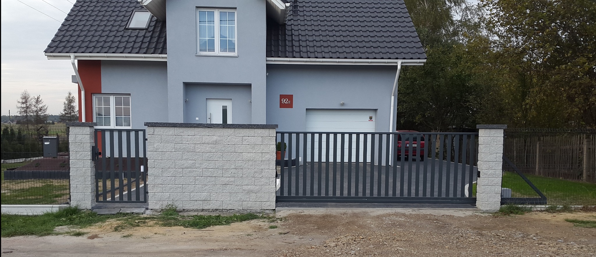 Nowoczesna, przesuwna brama wjazdowa z pionowymi, grafitowymi szczeblami, osadzona między słupkami z jasnoszarej kostki, na tle szarego domu z ciemnym dachem.