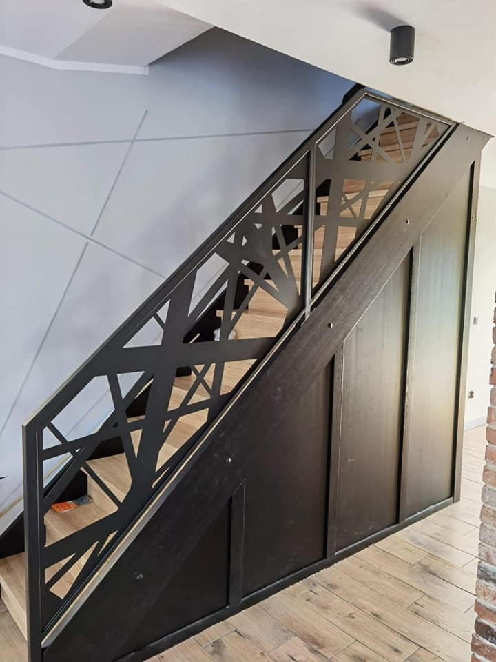 Nowoczesne schody z drewnianymi stopniami i czarną, ażurową balustradą o geometrycznym wzorze na tle szarej ściany z delikatnym graficznym motywem.