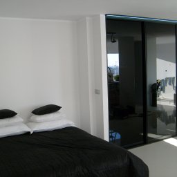 BUMEB RAFAŁ GUSTOŁEK - Nowoczesna sypialnia z czarno-białą pościelą i dużą szafą przesuwną z czarnymi szklanymi drzwiami, odbijającymi widok z balkonu.