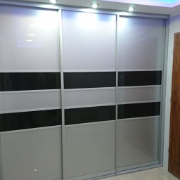 BUMEB RAFAŁ GUSTOŁEK - Trzydrzwiowa szafa przesuwna z białymi i czarnymi panelami, widoczna surowa ściana obok, prawdopodobnie w trakcie montażu.