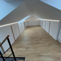 BUMEB RAFAŁ GUSTOŁEK - Adaptacja poddasza ze skosami: zabudowa z białych szaf z drewnianymi akcentami i oświetleniem LED, widok z góry schodów na drewnianą podłogę.