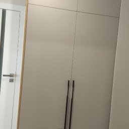 BUMEB RAFAŁ GUSTOŁEK - Jasnoszara szafa z trzema szufladami i długimi, czarnymi uchwytami, obok białe drzwi z drewnianą ramą. Minimalistyczny design, nowoczesne wykończenie.