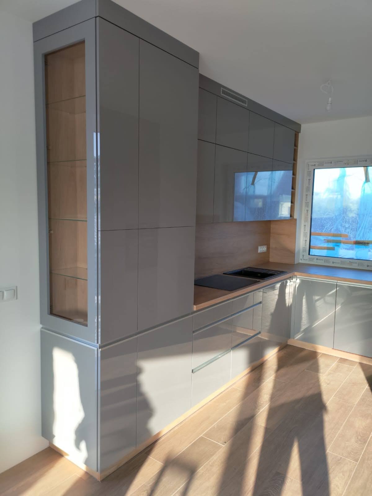 Nowoczesna, szara zabudowa kuchenna z przeszkloną witryną, drewnianym blatem i wbudowanym zlewem, oświetlona promieniami słonecznymi padającymi na podłogę.