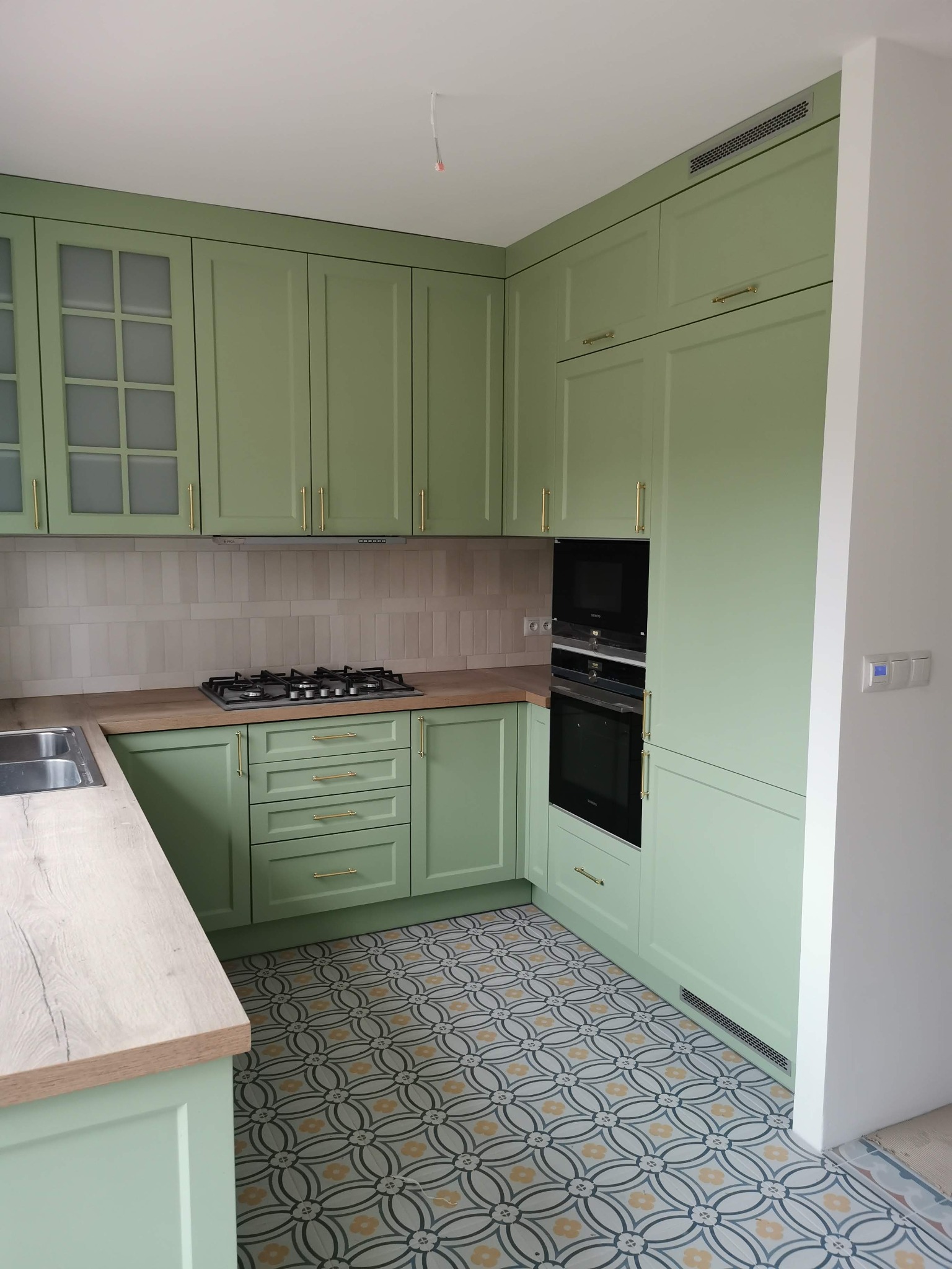 Jasnozielona zabudowa kuchenna z drewnianym blatem, płytą gazową i piekarnikiem w zabudowie, wzorzysta podłoga.