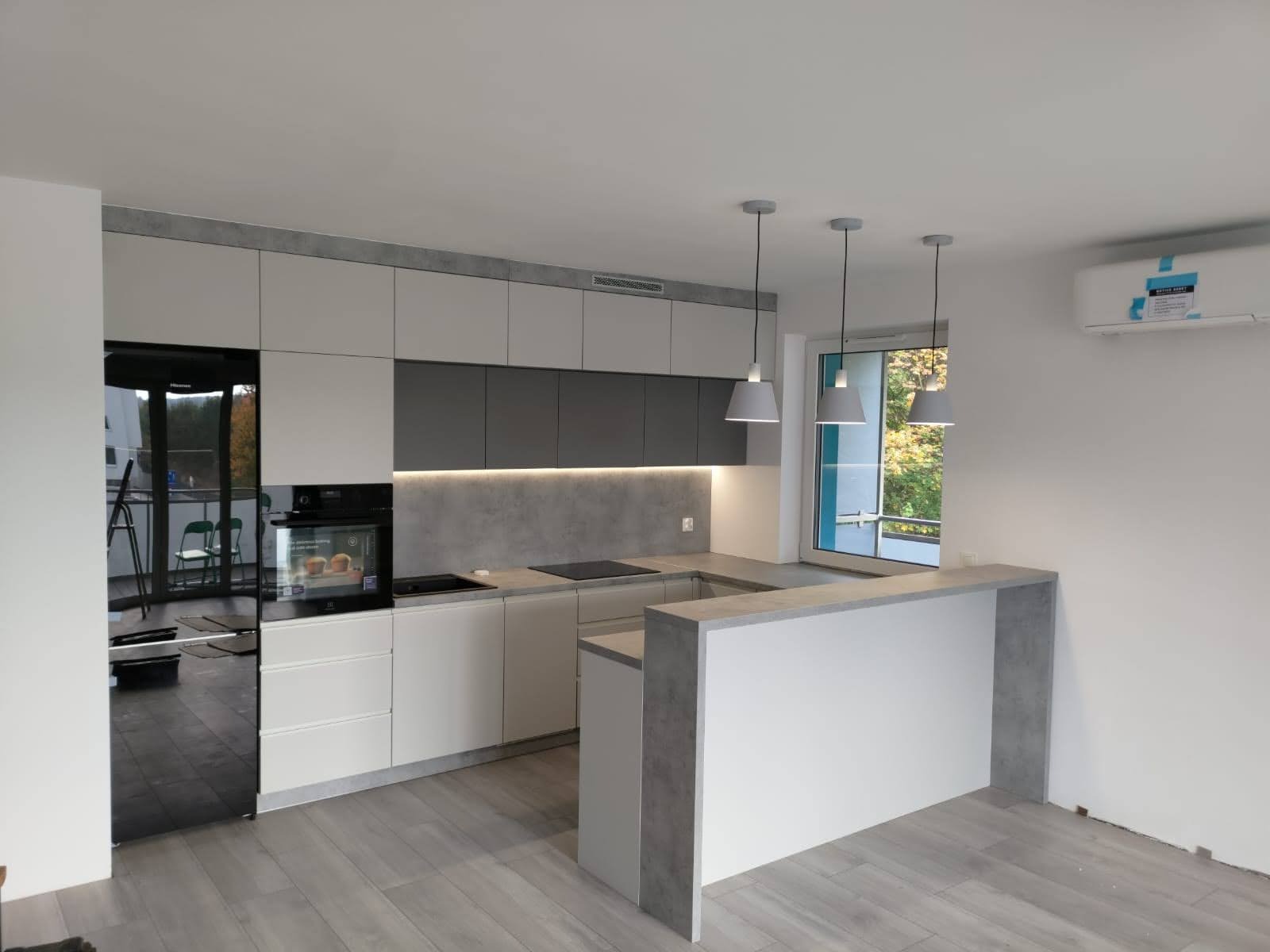 Nowoczesna kuchnia z białymi i szarymi szafkami, czarną lodówką i blatem w kolorze betonu, oświetlona trzema wiszącymi lampami, z widokiem na zieleń za oknem.