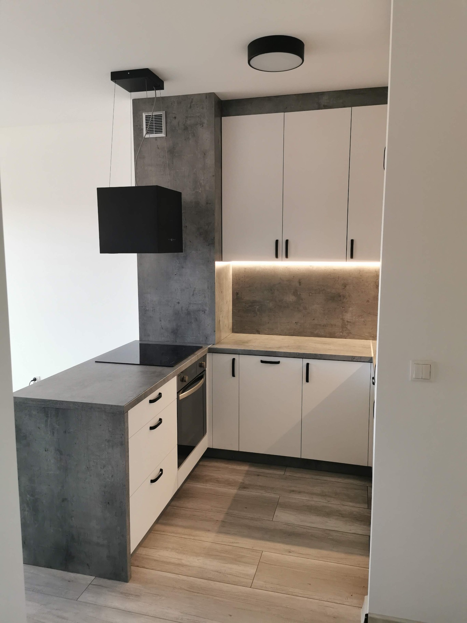 Nowoczesna kuchnia narożna z białymi szafkami i blatem imitującym beton, oświetlenie LED podszafkowe, czarny okap i płyta indukcyjna.