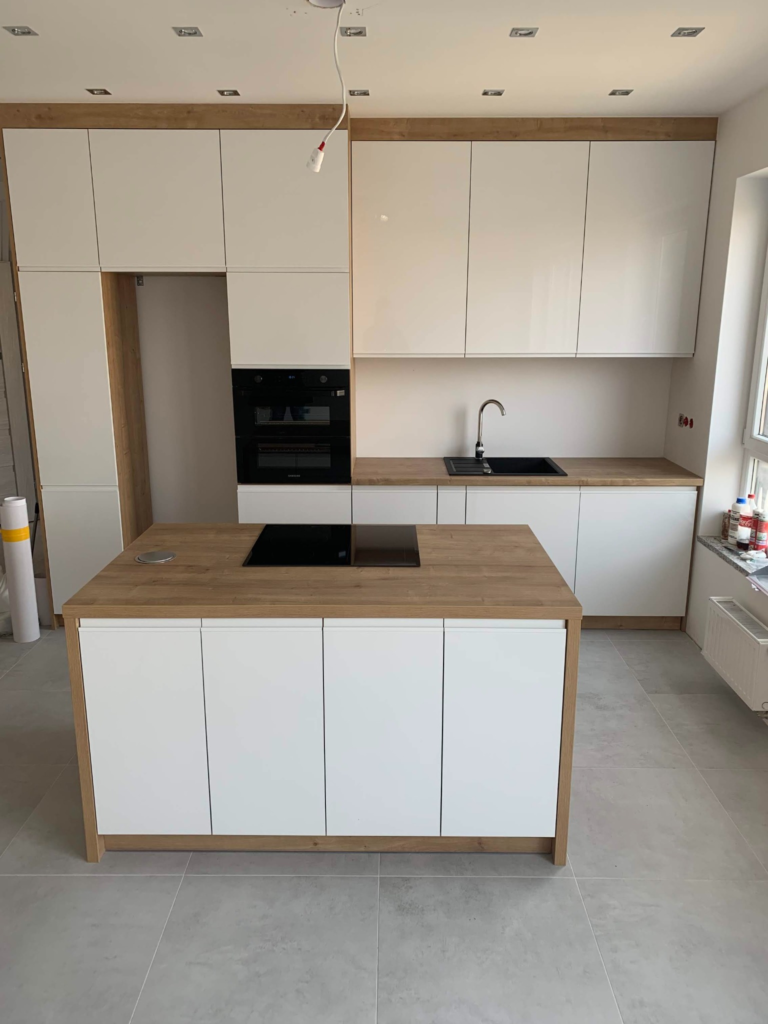 Nowoczesna kuchnia z wyspą, białe fronty szafek z drewnianym blatem, czarna płyta indukcyjna i zlewozmywak, widoczne niedokończone prace instalacyjne.