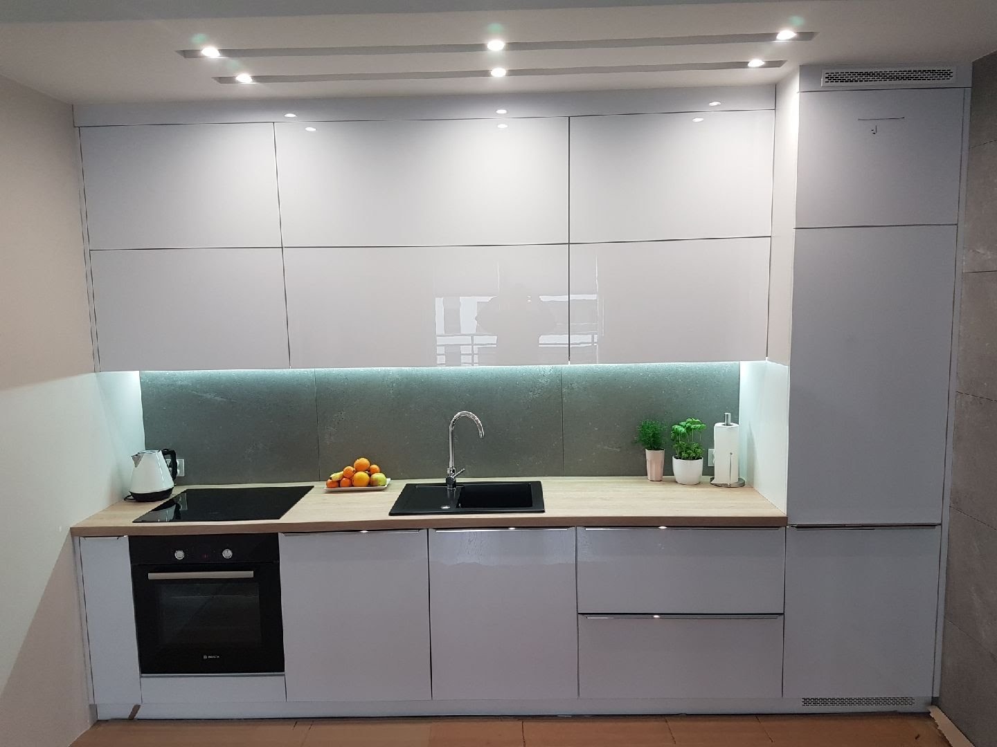 Nowoczesna, minimalistyczna kuchnia z białymi, lakierowanymi frontami, drewnianym blatem i szarym panelem ściennym. Widoczny czarny zlew, płyta indukcyjna, piekarnik, owoce w misce i małe rośliny...