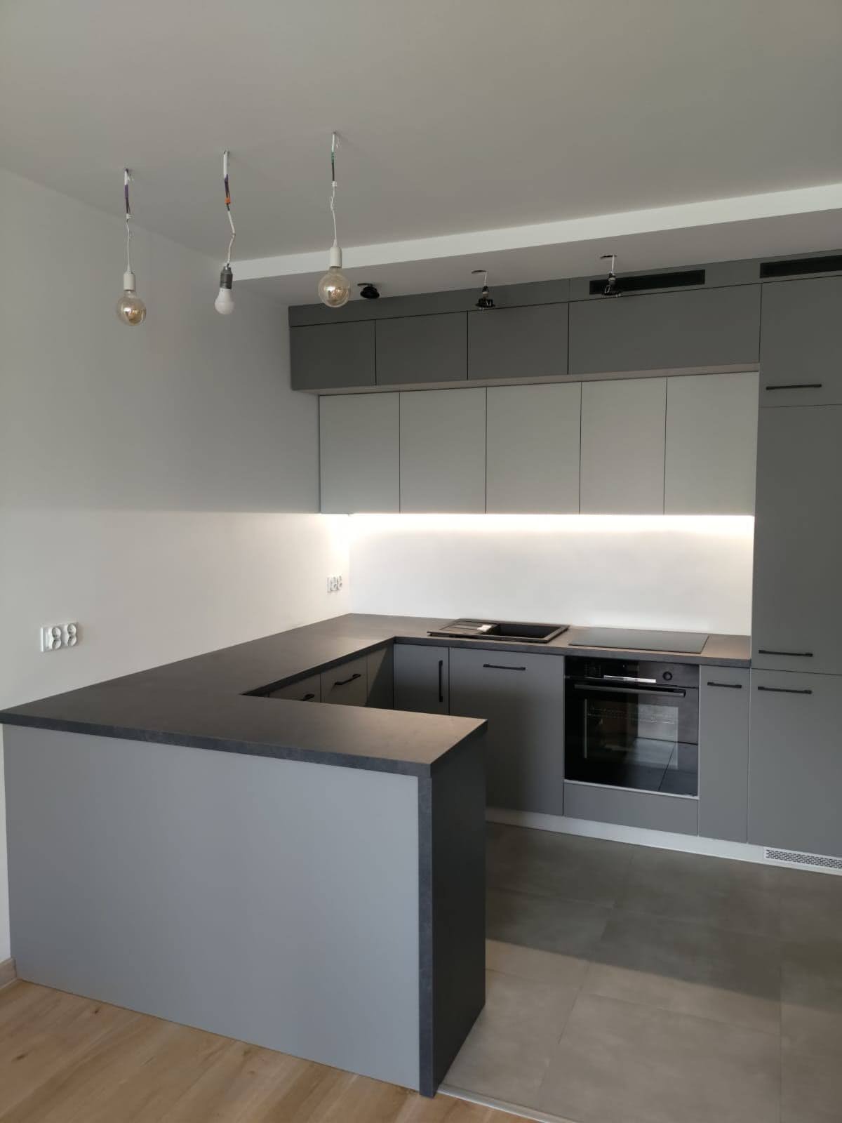 Nowoczesna, szara kuchnia z wyspą i oświetleniem LED pod szafkami górnymi, z trzema wiszącymi żarówkami bez kloszy.