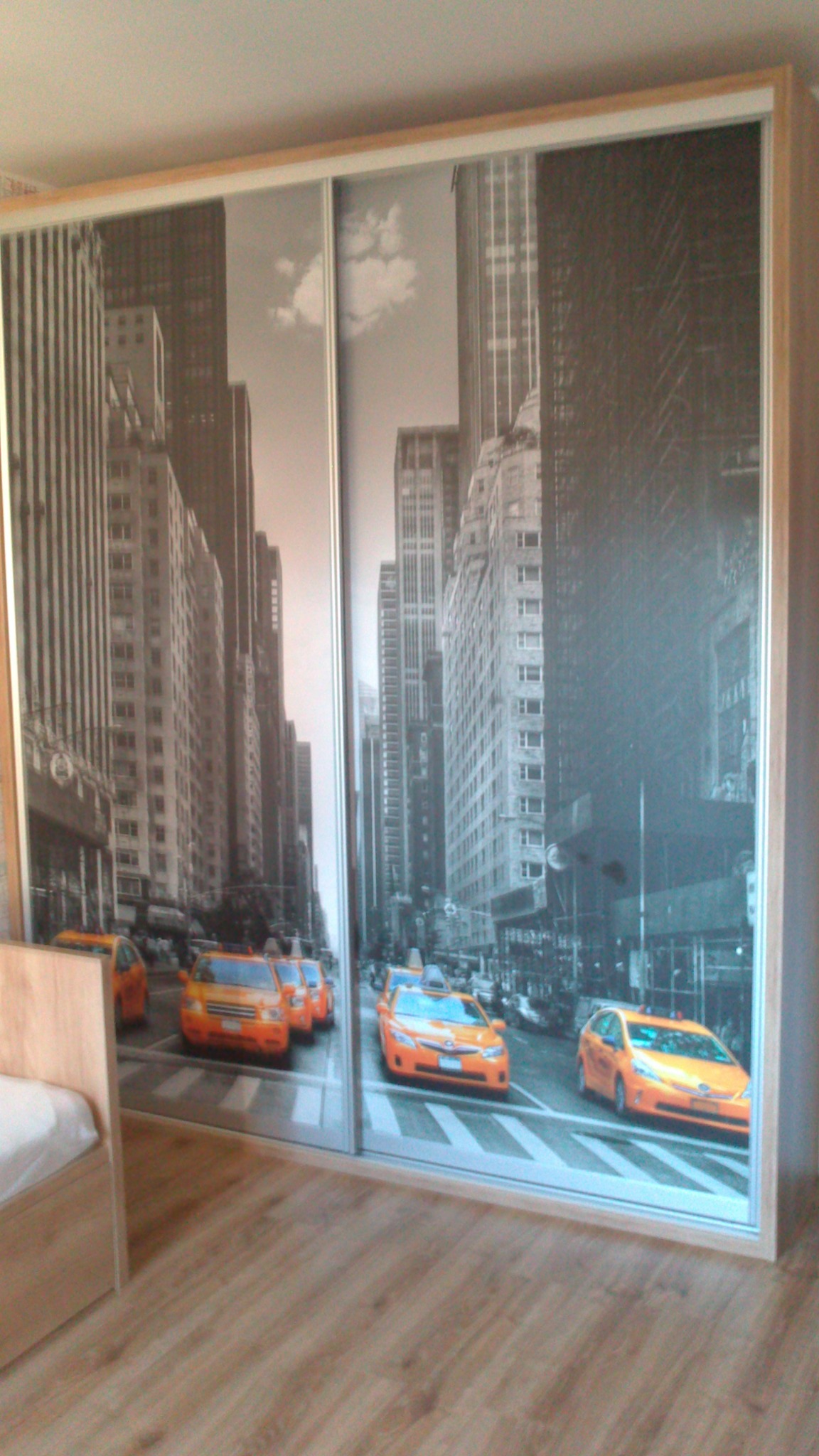 Szafa przesuwna z fototapetą przedstawiającą ulicę w Nowym Jorku z żółtymi taksówkami, widoczna część łóżka obok.