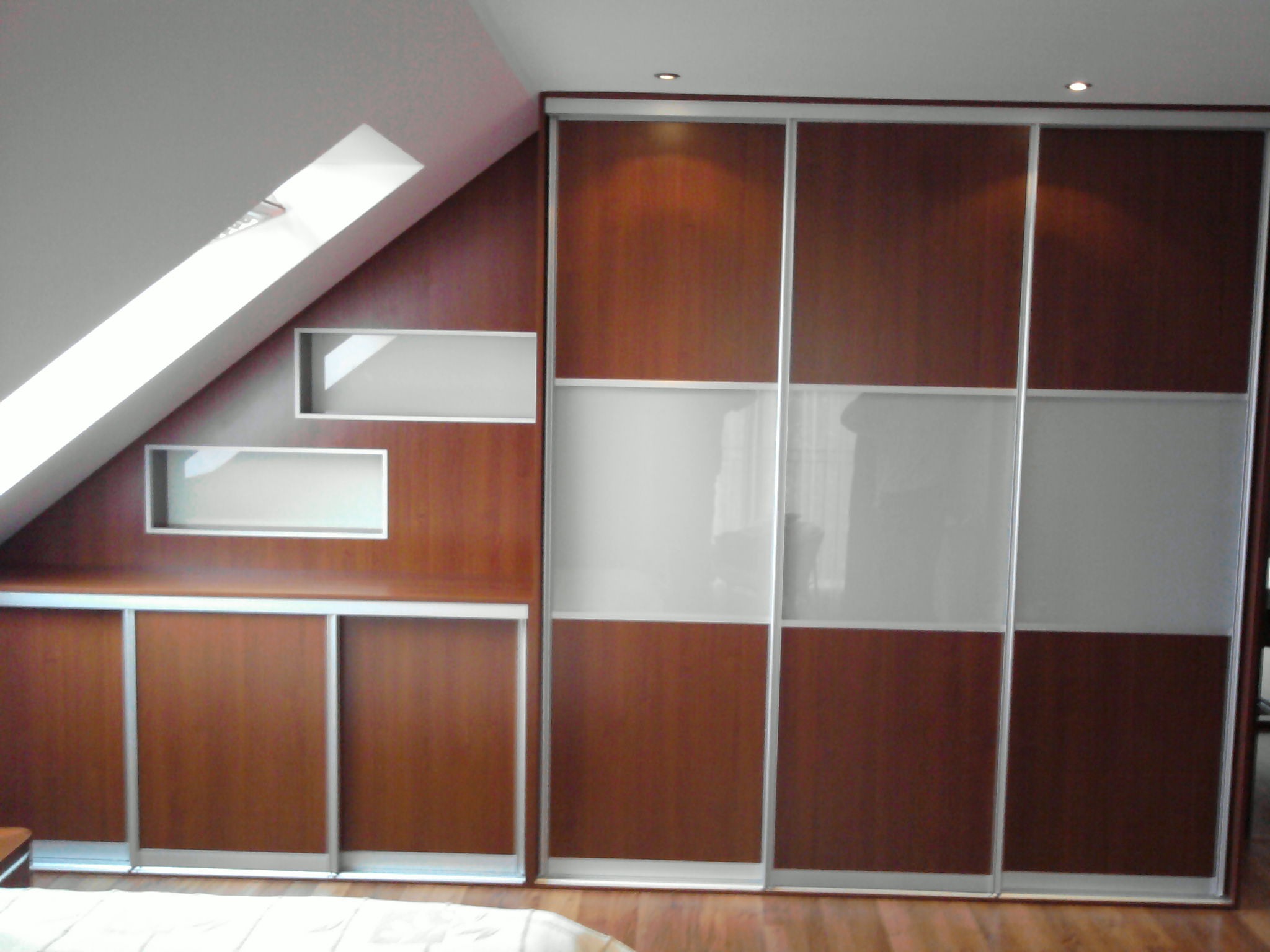 Szafa wnękowa z przesuwnymi drzwiami w kolorze brązowym i białym, dopasowana do skosu poddasza, z dodatkowymi półkami wbudowanymi w ścianę.