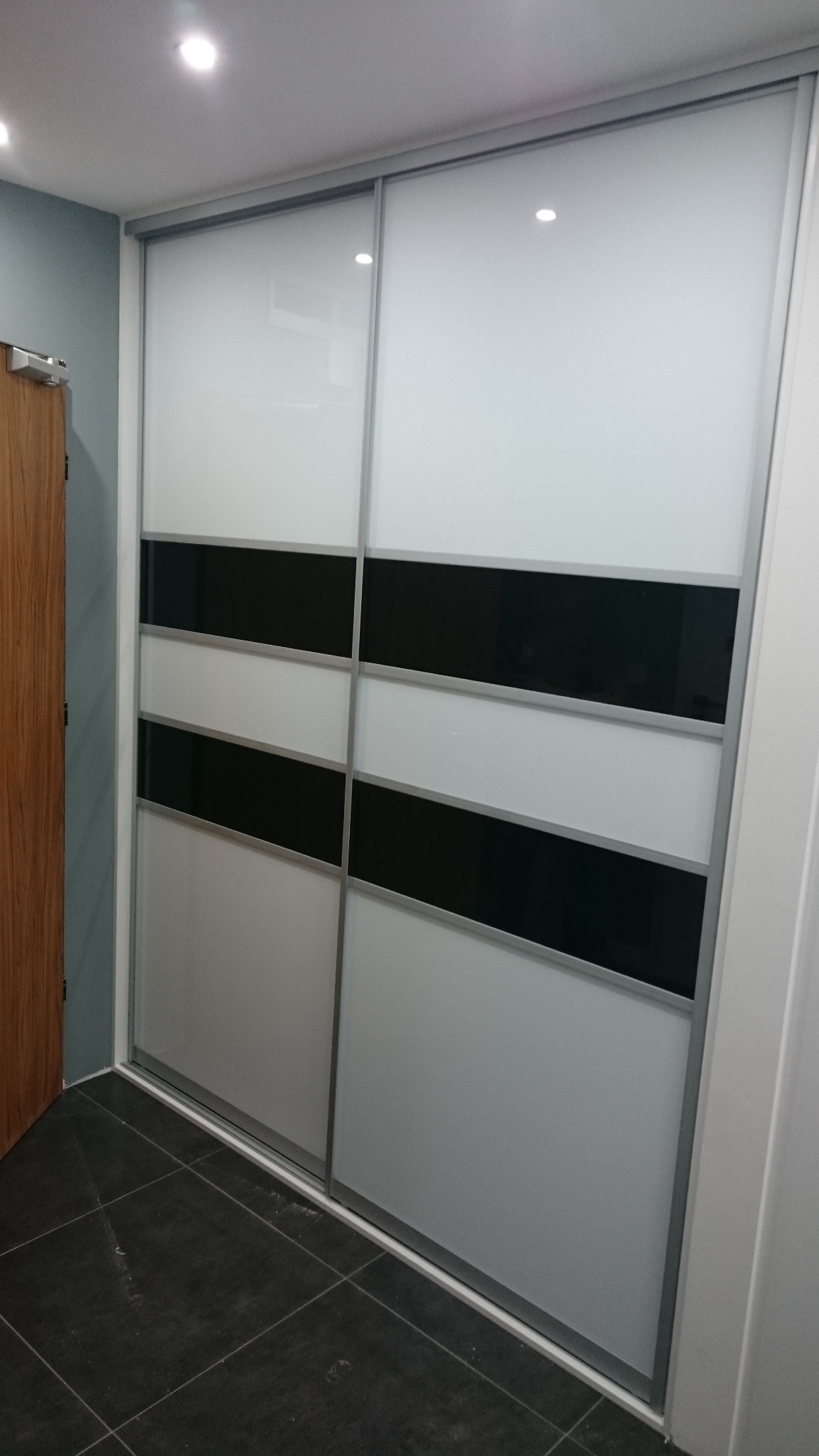 Nowoczesna szafa wnękowa z przesuwnymi drzwiami, biało-czarne poziome pasy, srebrne ramy, widoczna część podłogi z ciemnych płytek.