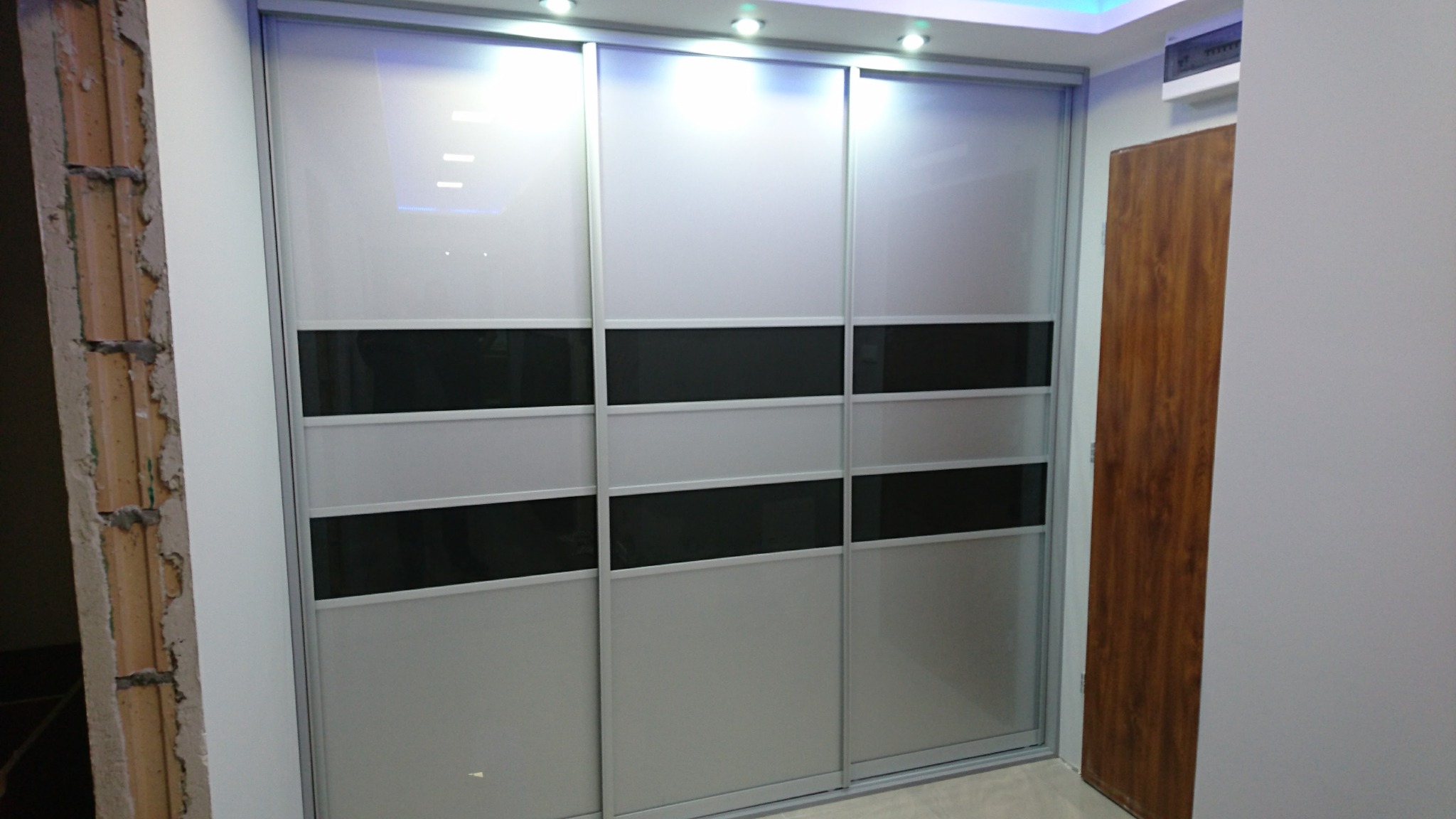 Trzydrzwiowa szafa przesuwna z białymi i czarnymi panelami, widoczna surowa ściana obok, prawdopodobnie w trakcie montażu.