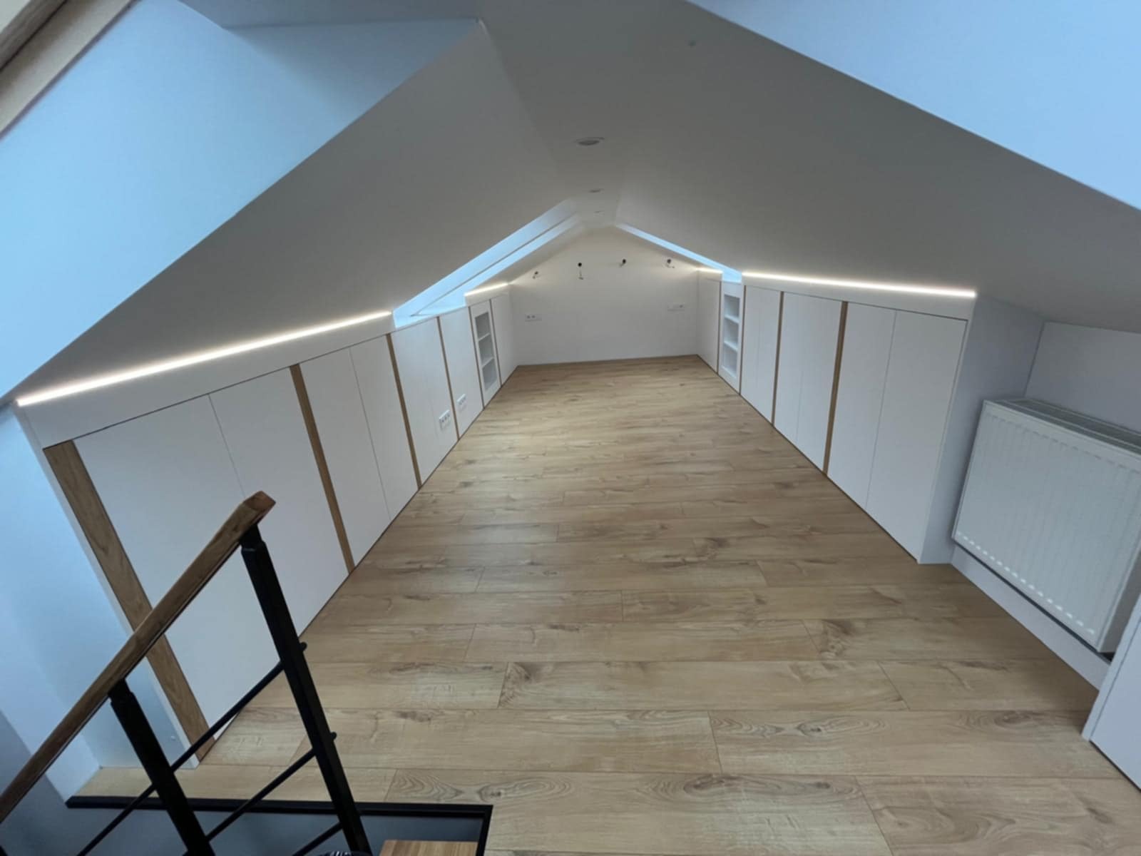 Adaptacja poddasza ze skosami: zabudowa z białych szaf z drewnianymi akcentami i oświetleniem LED, widok z góry schodów na drewnianą podłogę.