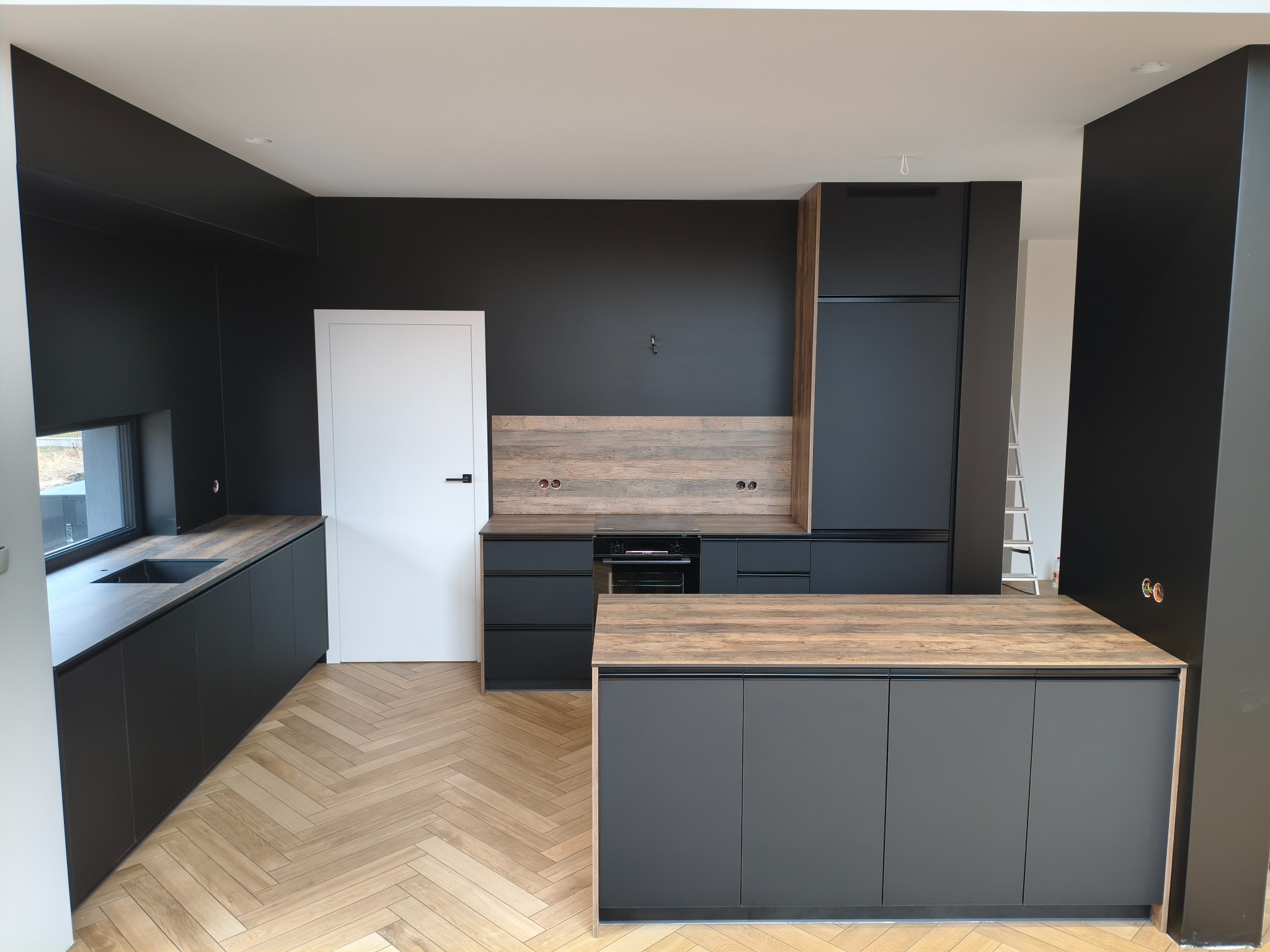 Nowoczesna kuchnia z czarnymi szafkami i drewnianym blatem, parkiet w jodełkę, minimalistyczny design, oświetlenie punktowe, widok na okno i białe drzwi.