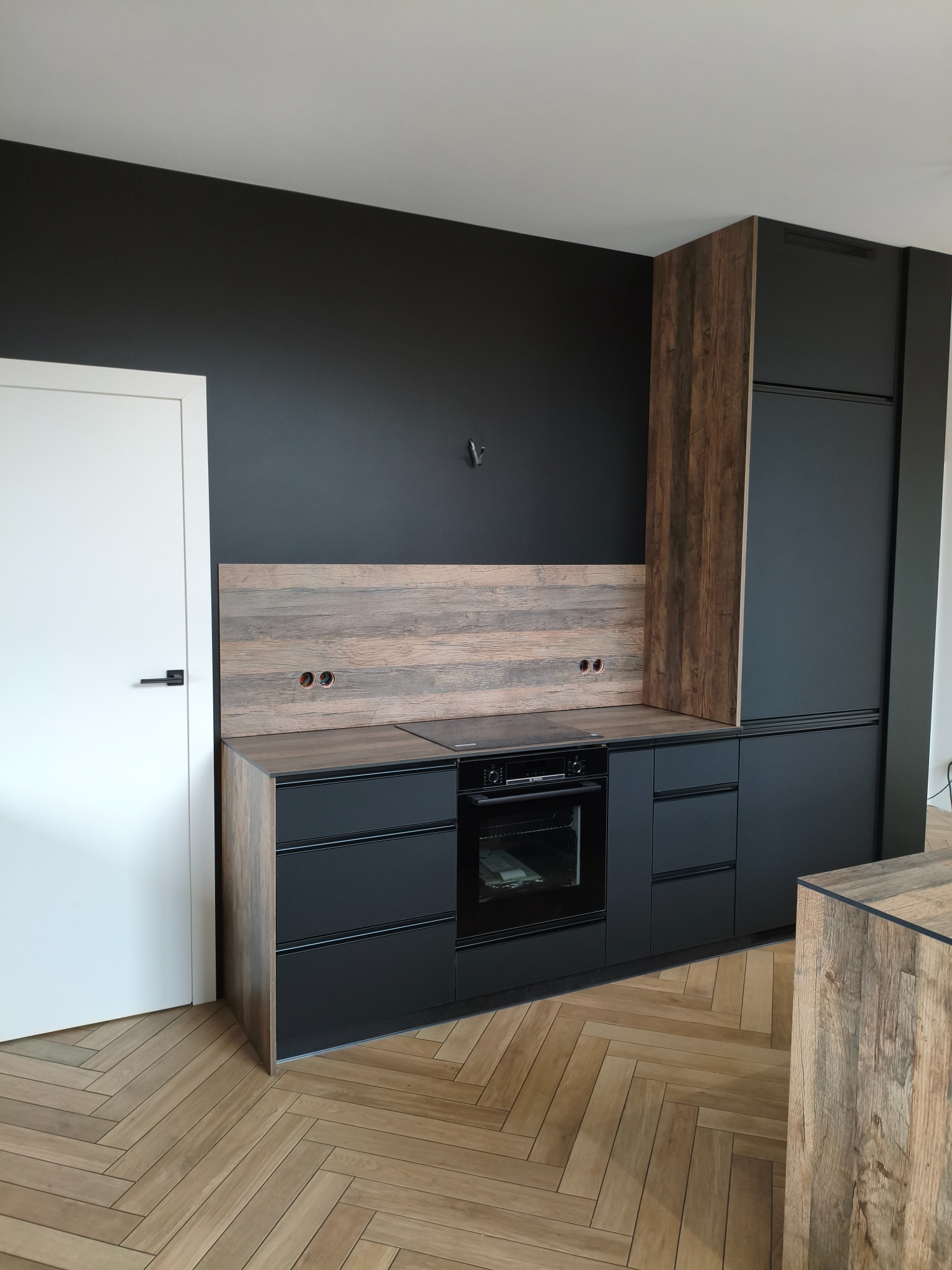 Nowoczesna kuchnia z czarnymi frontami i drewnianymi akcentami, widok na zabudowę z piekarnikiem i lodówką, parkiet w jodełkę, czarna ściana.