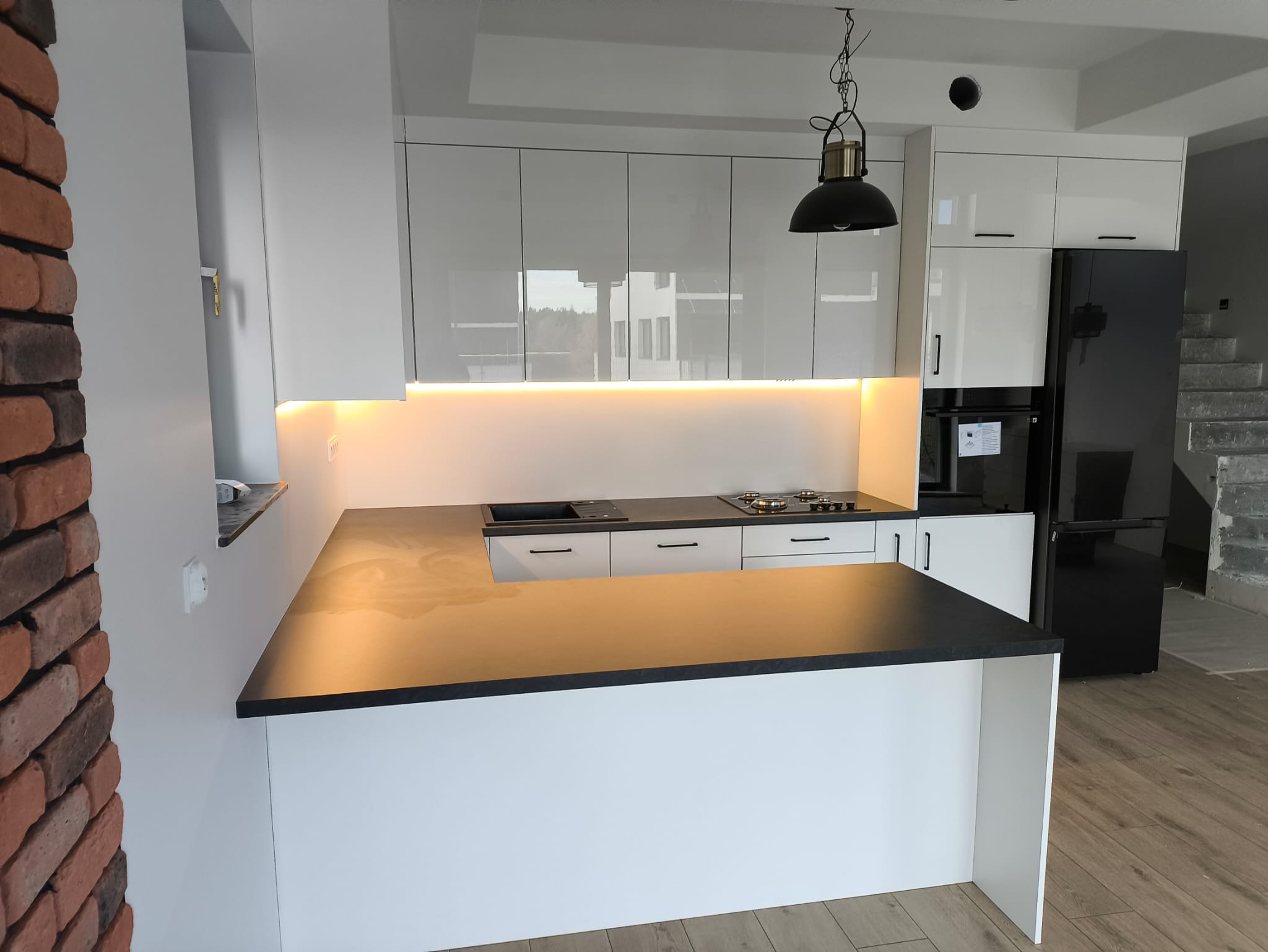 Nowoczesna kuchnia z białymi, lakierowanymi frontami, czarnym blatem i ceglaną ścianą. Oświetlenie LED pod szafkami górnymi. Widoczny fragment wyspy kuchennej.