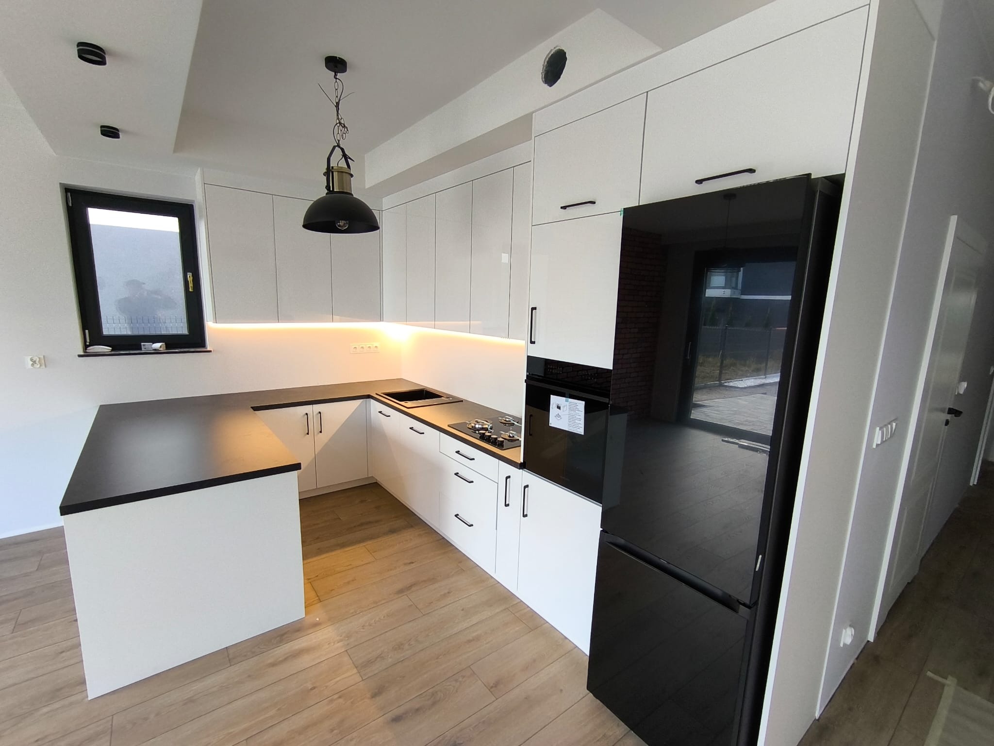 Nowoczesna kuchnia z białymi szafkami i czarnym blatem, oświetlona lampą wiszącą. Widoczny sprzęt AGD w czarnym kolorze i drewniana podłoga.
