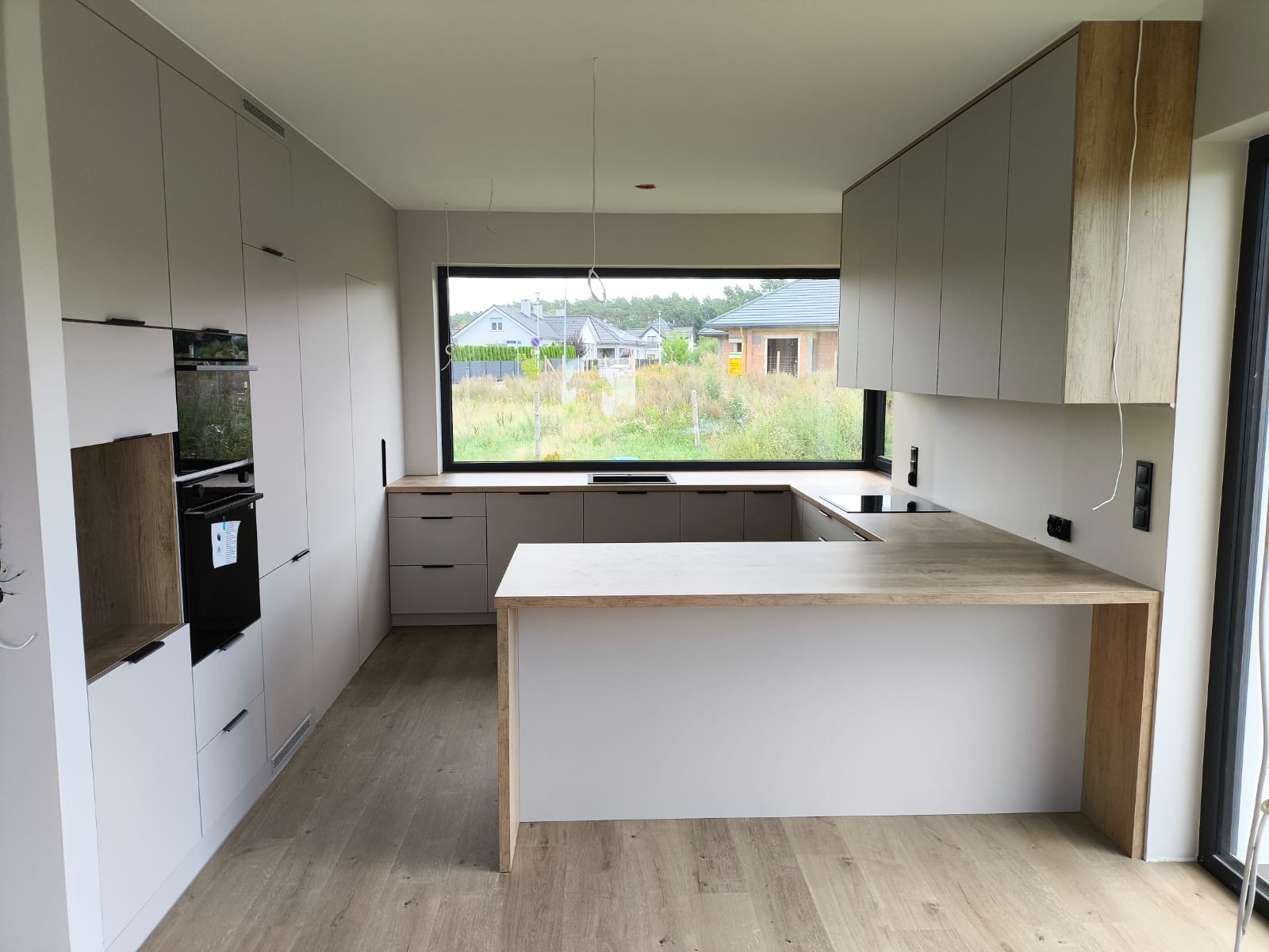 Jasna kuchnia z wyspą i dużym oknem z widokiem na zieleń. Minimalistyczny design, drewniany blat, nowoczesne oświetlenie. Widoczne gniazdka elektryczne na ścianie.