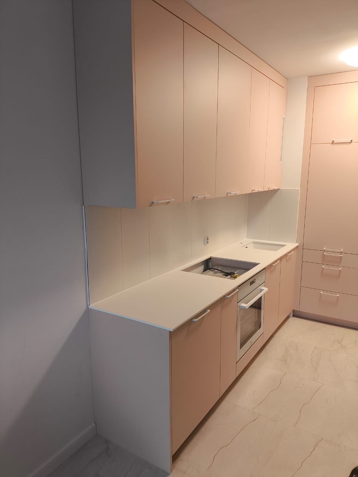 Jasnoróżowa zabudowa kuchenna z białym blatem i sprzętem AGD. Minimalistyczny design, widok na szafki wiszące i stojące, marmurowa podłoga.