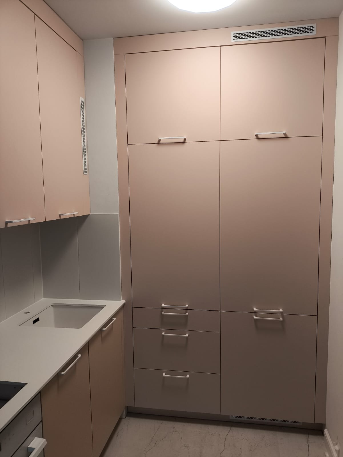 Jasna, minimalistyczna kuchnia z zabudową po sufit. Fronty szafek w odcieniach beżu i szarości, z prostymi, podłużnymi uchwytami. Blat w kolorze złamanej bieli.