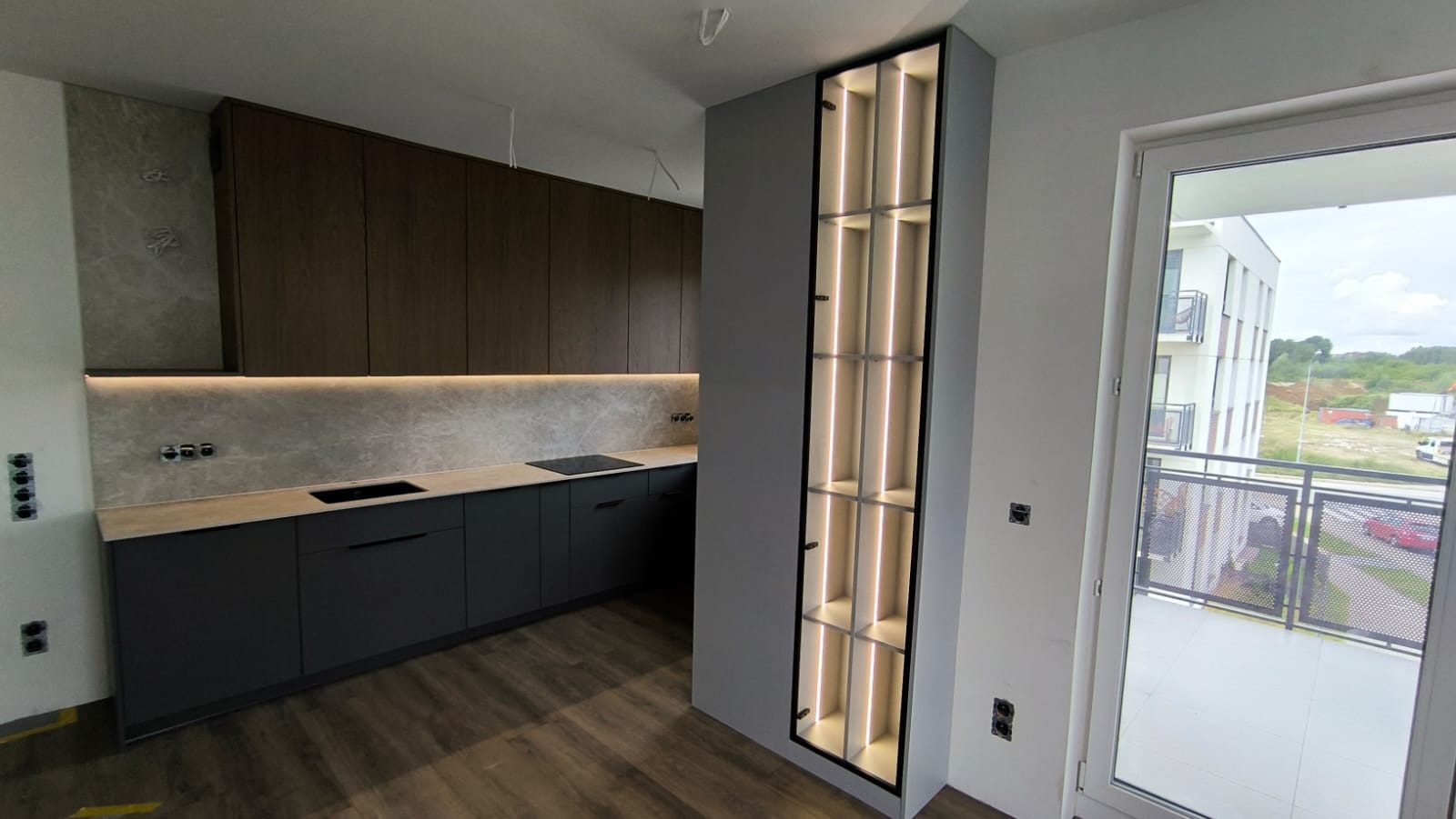 Nowoczesna kuchnia z ciemnymi szafkami i podświetlaną wnęką, widok na balkon i budynek w tle. Minimalistyczny design, drewniana podłoga.